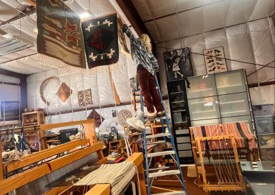 Cluttered workshop with wooden tables, shelves, ladders, and hanging fabric under a high ceiling