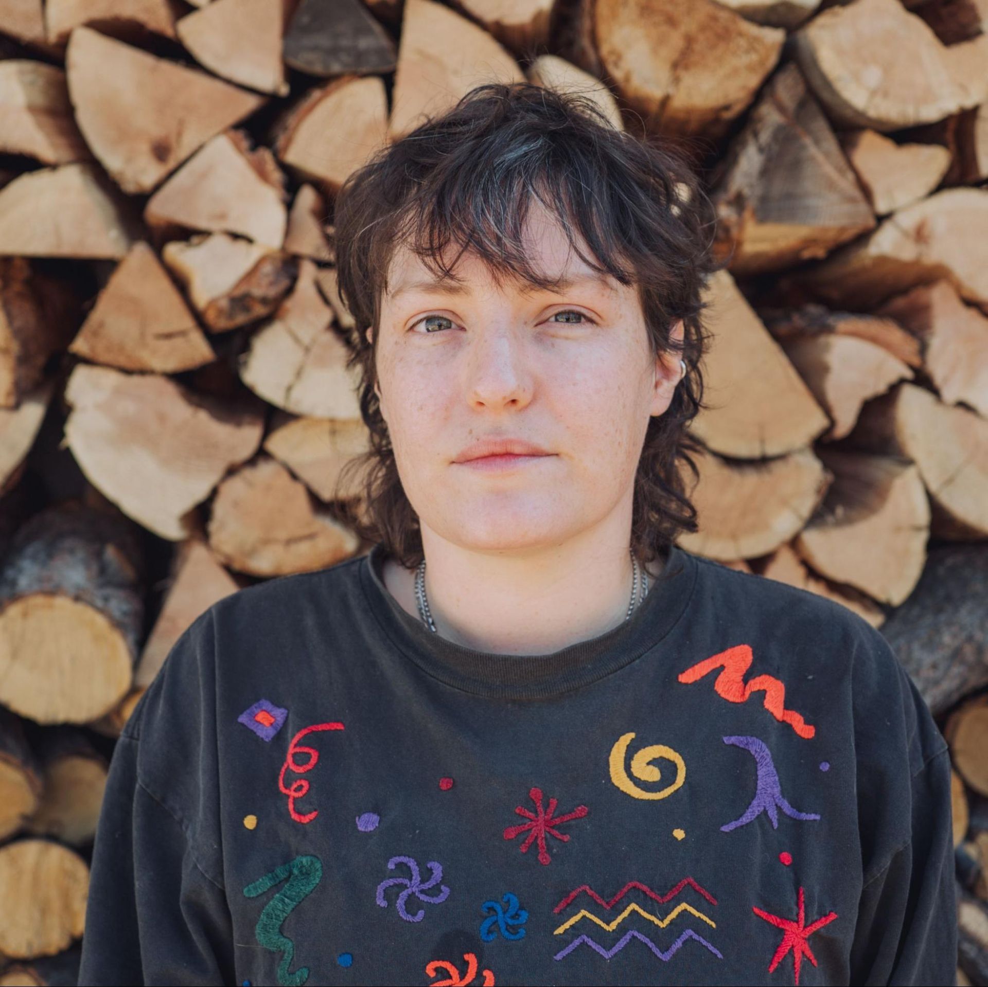 Person in a dark embroidered shirt standing before stacked firewood.