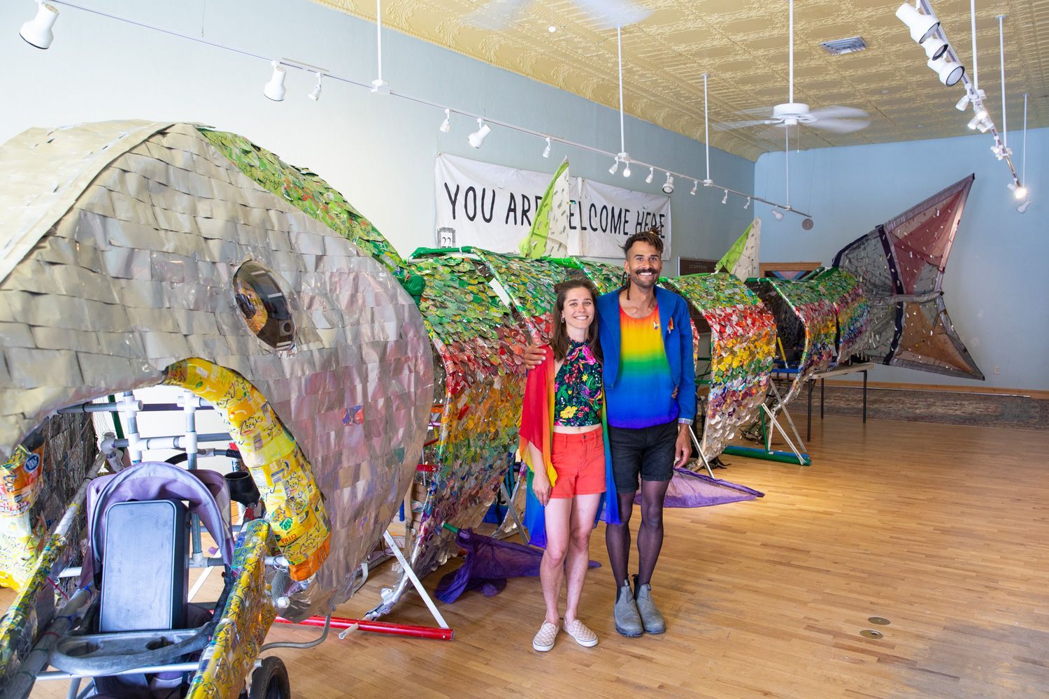 Two people stand beside a giant colorful fish sculpture in a bright gallery space.