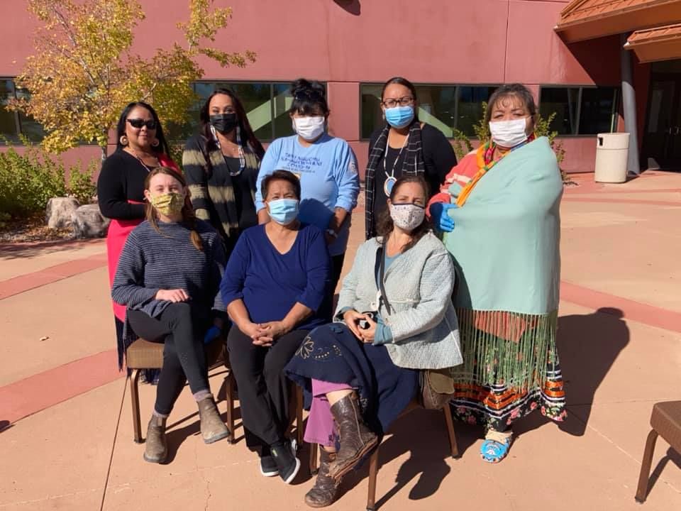 Seven masked people posing outdoors in front of a pink building, some seated and some standing.