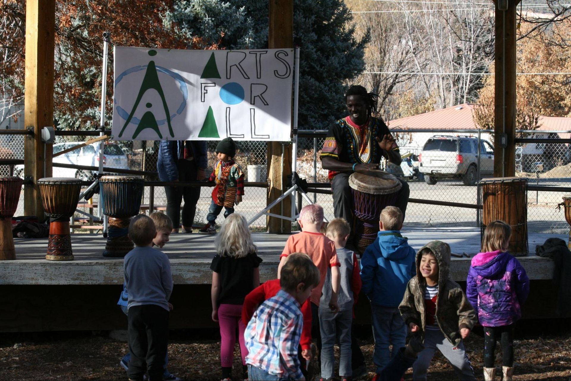 Outdoor stage performance with children watching in front of a banner reading “Arts for …”