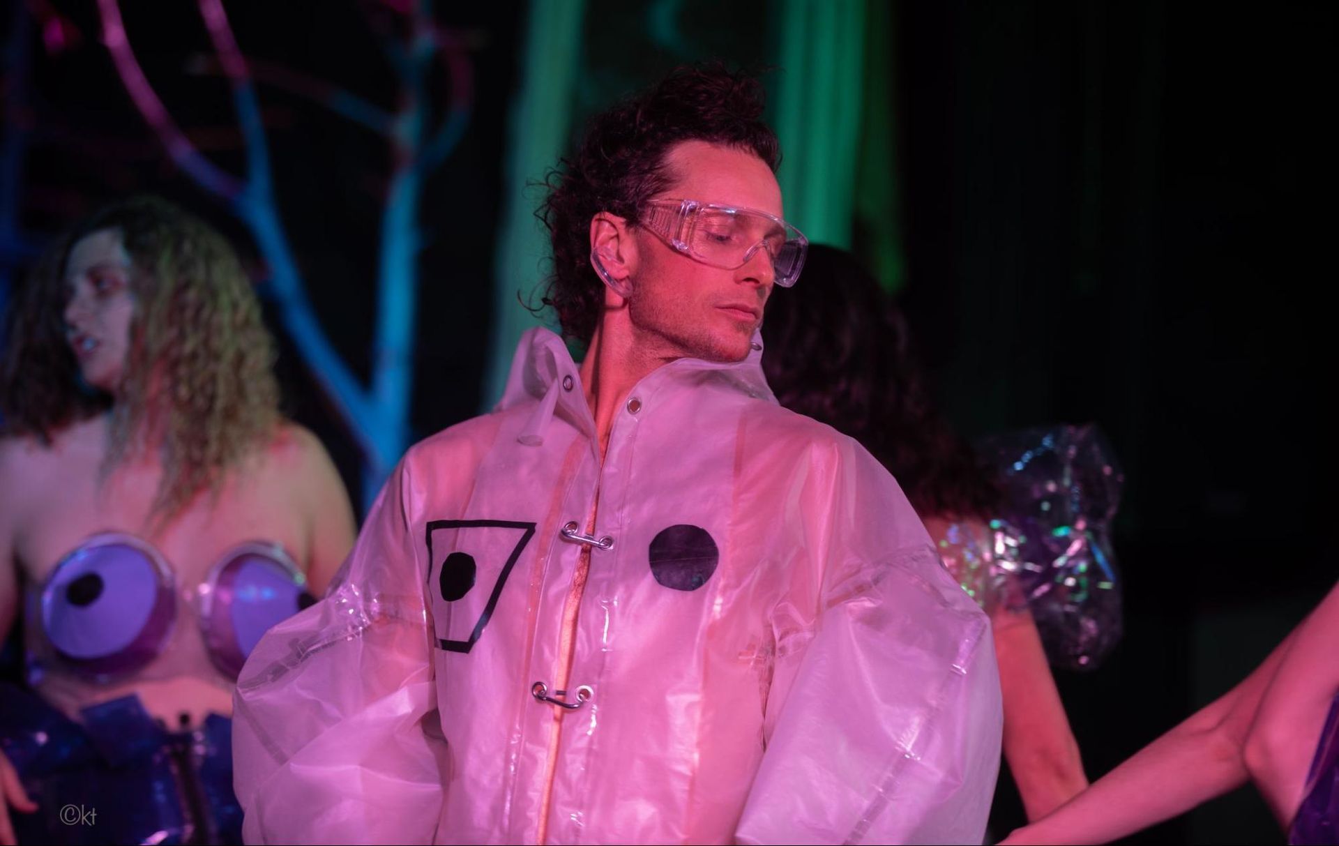 Person in a clear raincoat and glasses stands under colorful stage lights with performers in the background