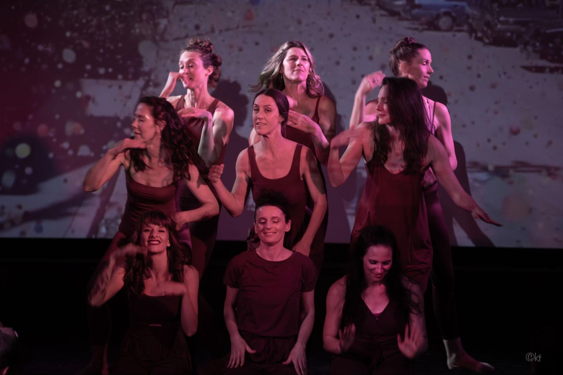 Ensemble of dancers in dark red lighting performing onstage against a patterned backdrop.