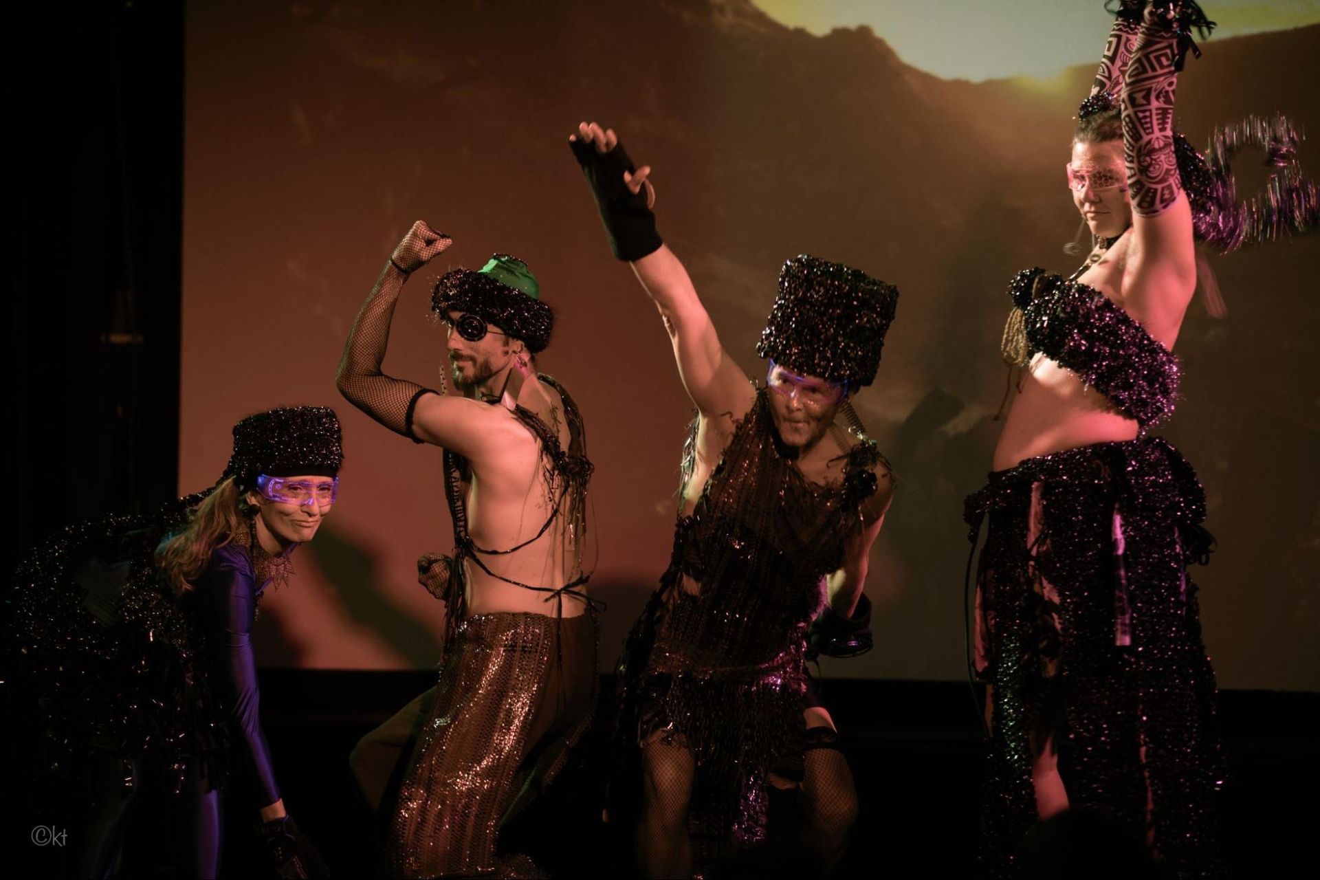 Four dancers in sequined costumes perform onstage under warm pink and orange lights.