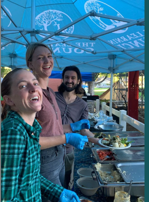 Three people smiling and serving food under a blue canopy at an outdoor buffet