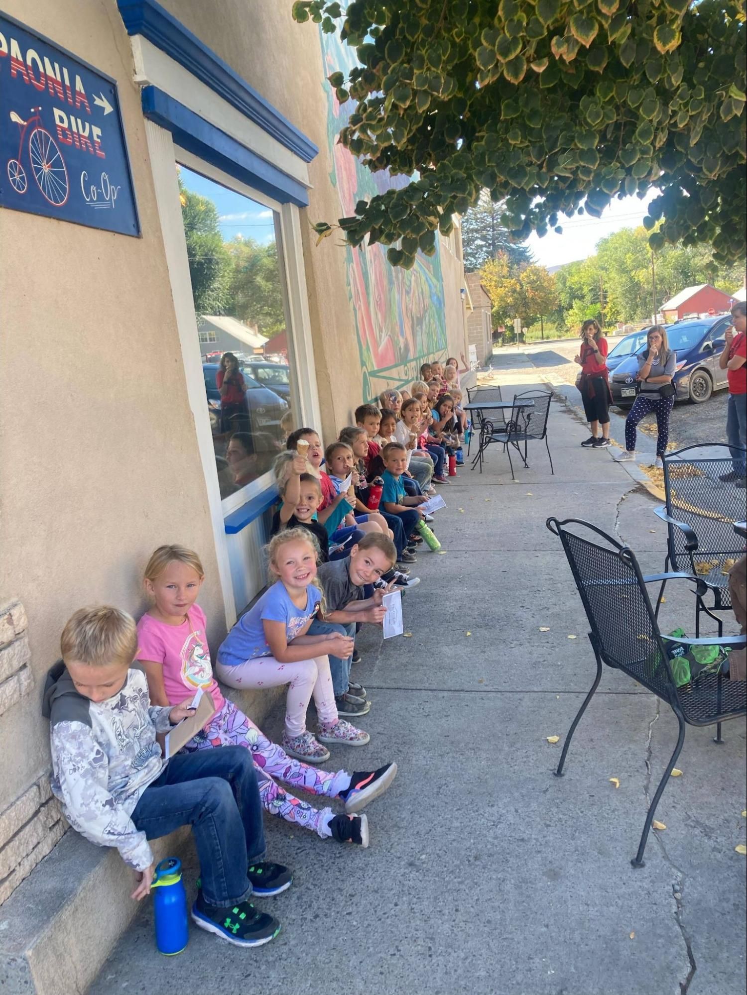 Children sitting along a shaded sidewalk outside a café, with chairs and people in the background.