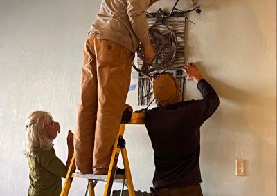 Three workers install wiring on a wall ladder in a room with a bicycle hanging nearby.