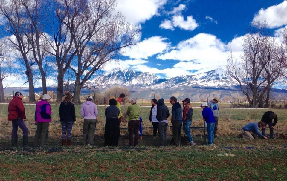 People standing outdoors by a stone wall, with snowy mountains and blue sky in the background.