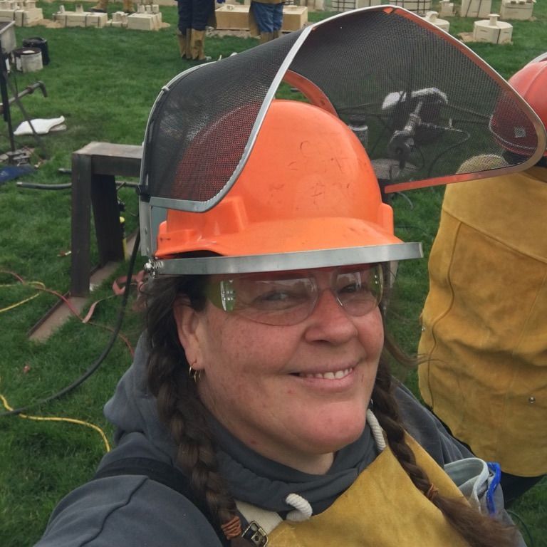 Person smiling outdoors in orange hard hat with face shield, wearing safety glasses at a worksite.