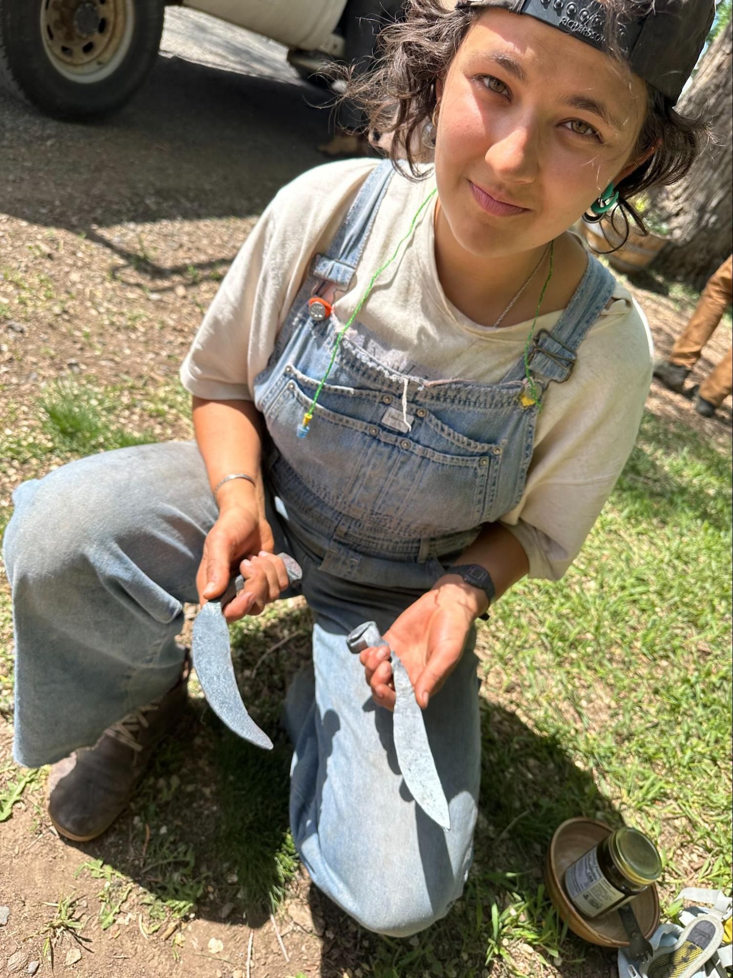 Person in denim overalls holding two fish outdoors on grass, with a vehicle in the background