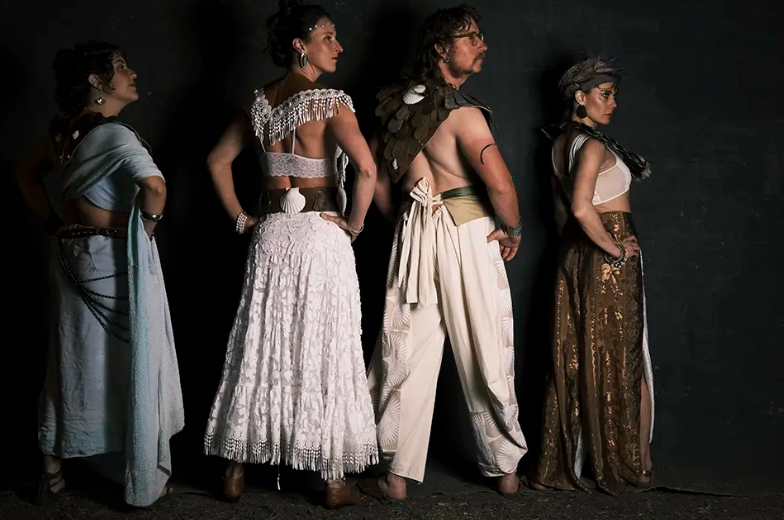 Four performers in ornate costumes pose against a dark backdrop, facing away from the camera.