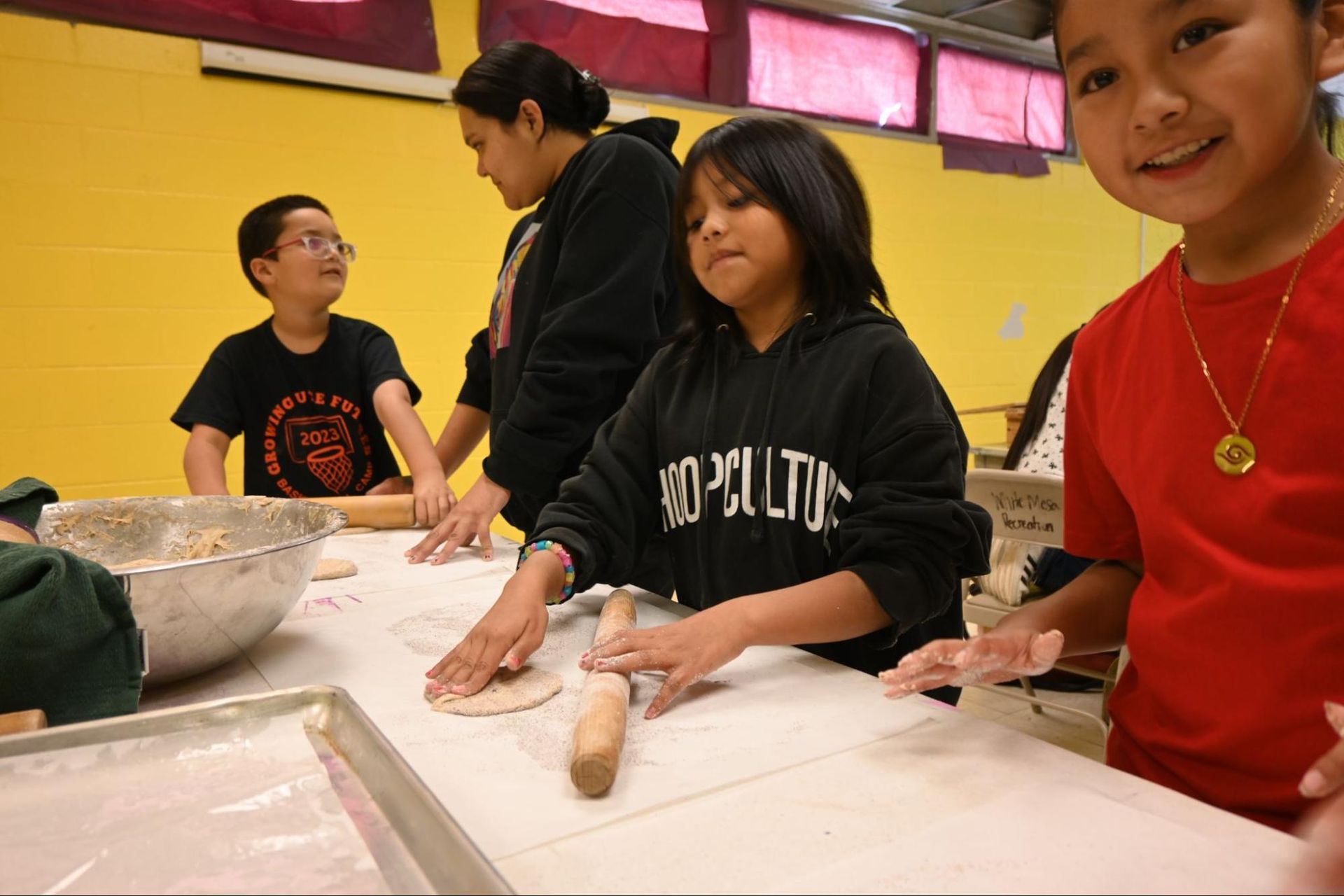 People rolling dough at a table in a bright kitchen or classroom, with a bowl of flour nearby.