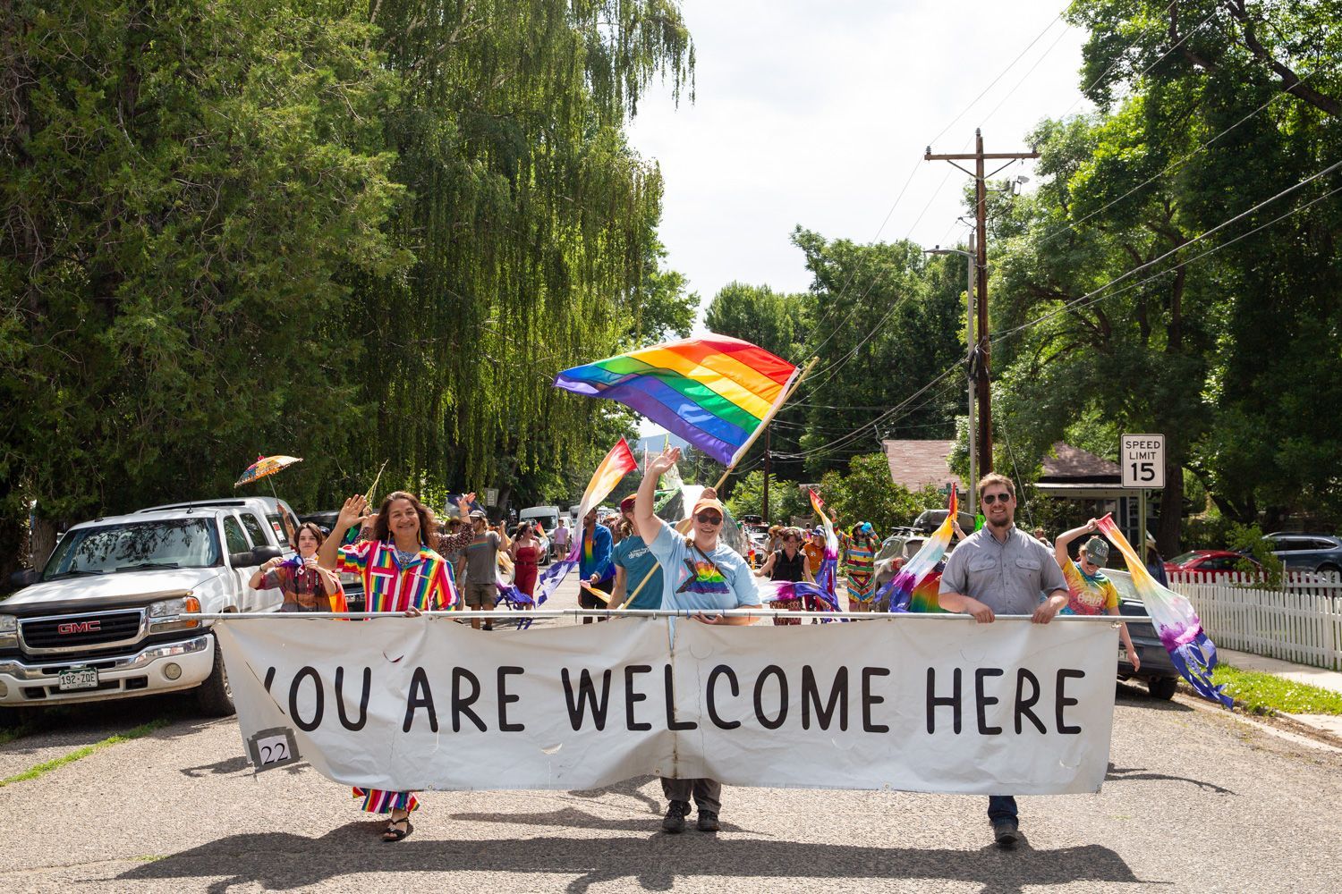 Pride parade marchers hold a “YOU ARE WELCOME HERE” banner with a rainbow flag on a tree-lined street