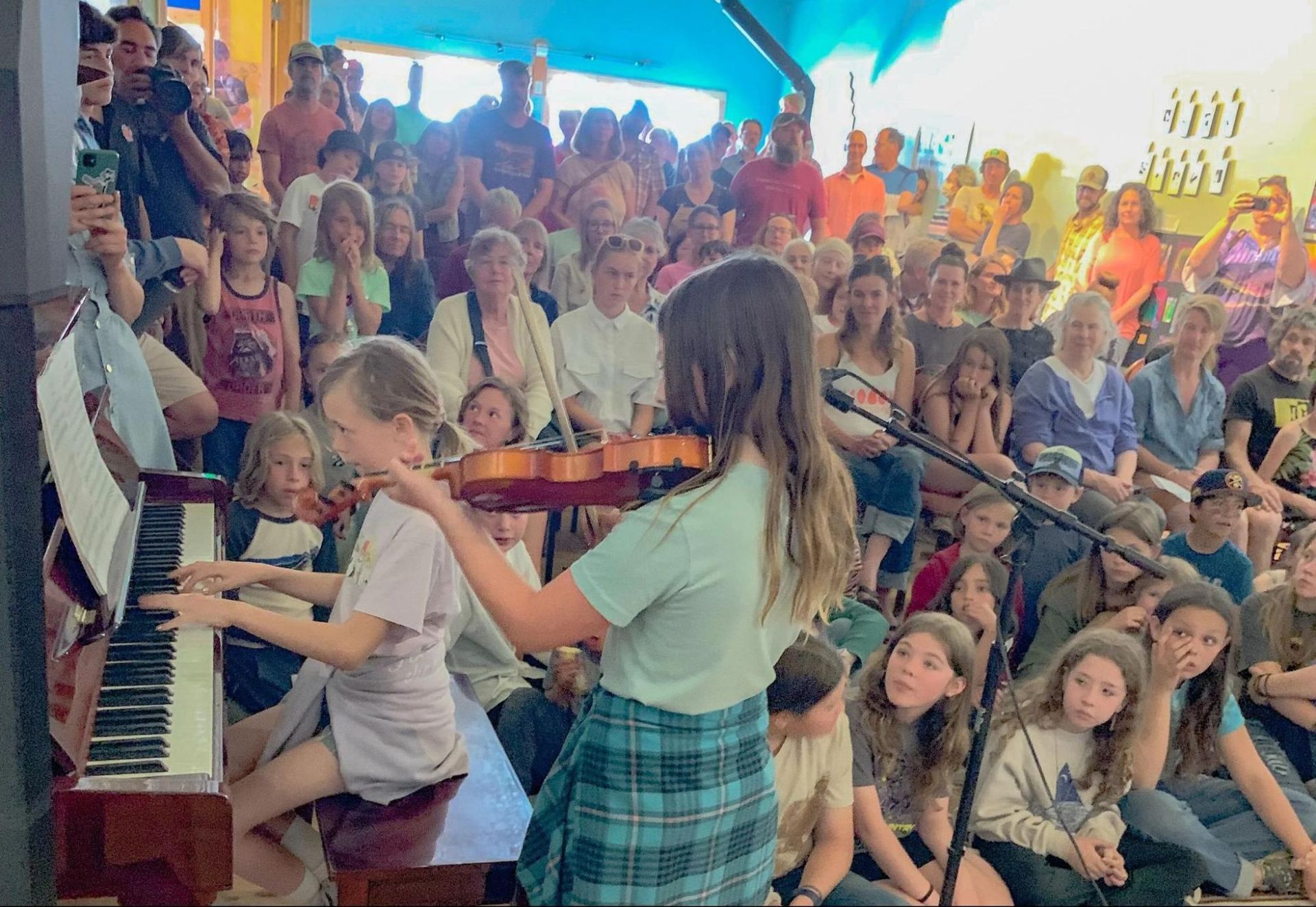 Child playing piano and violin for a seated crowd in a bright indoor venue