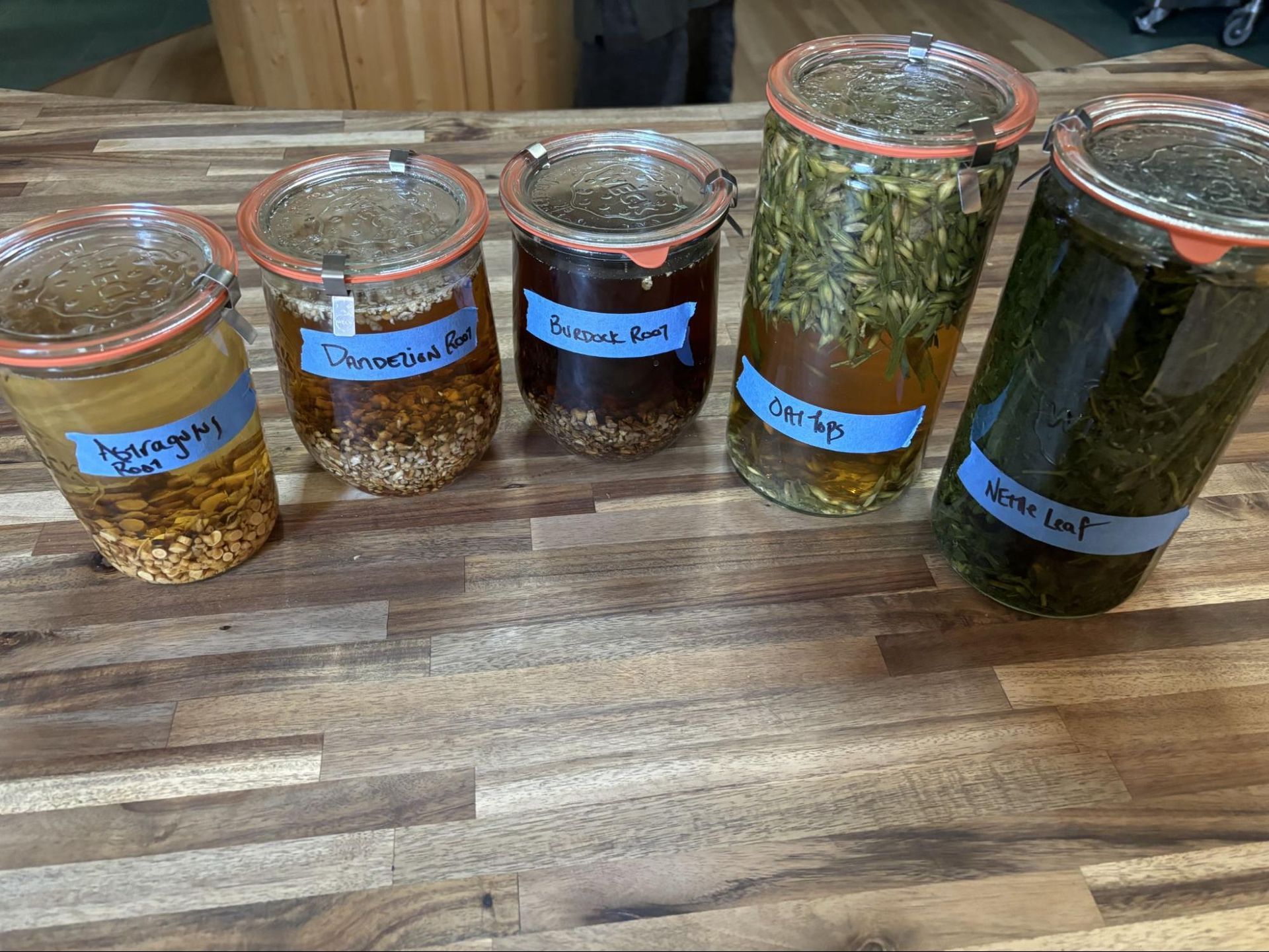 Five glass jars of dried herbs and spices lined up on a wooden table with handwritten labels.