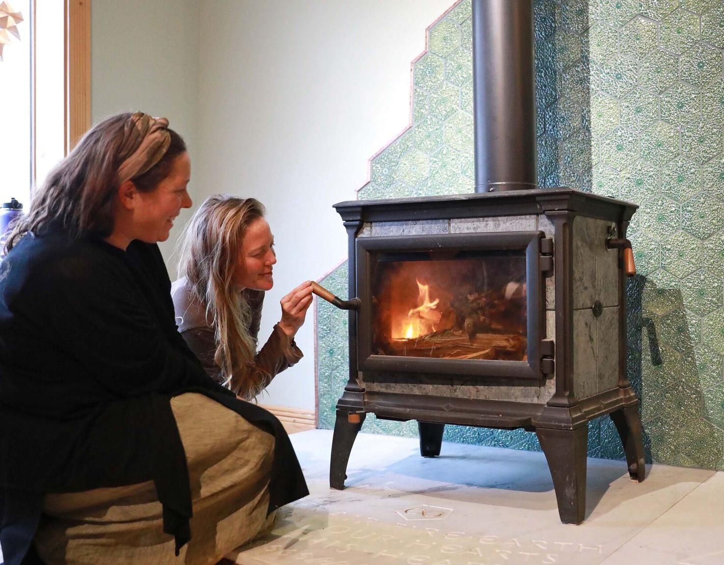 Two people kneel beside a lit wood stove in a room with a green mosaic wall.