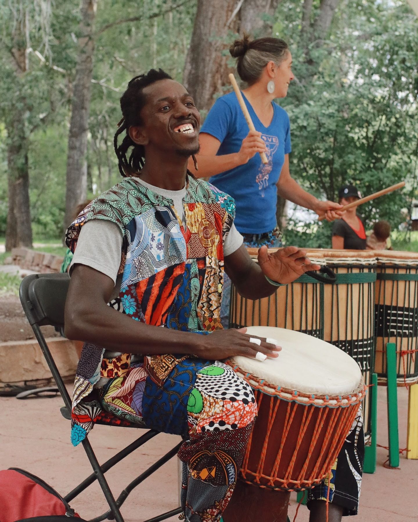 Drummer playing a hand drum outdoors while a person stands behind him near percussion instruments.