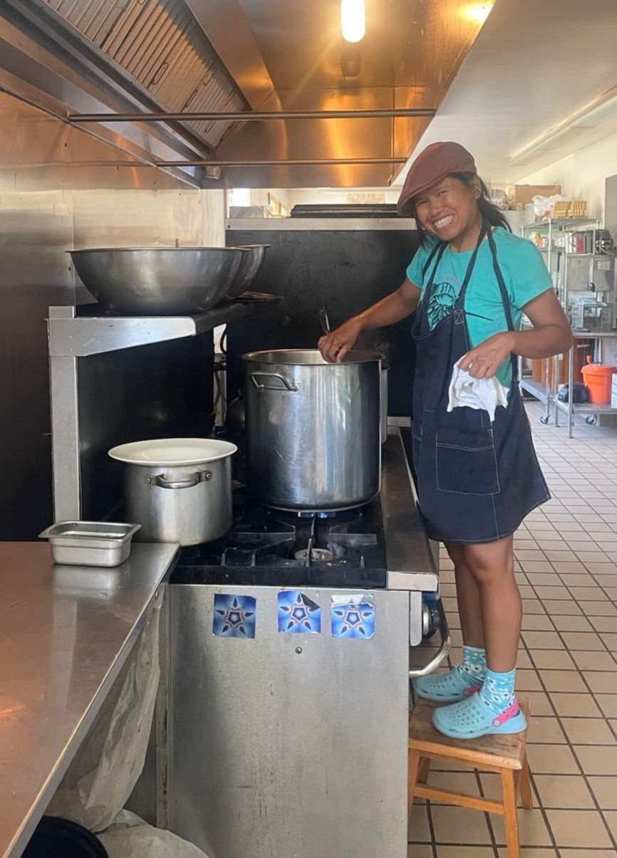 Person in a kitchen stirring a large pot on a stove, wearing an apron and headscarf.