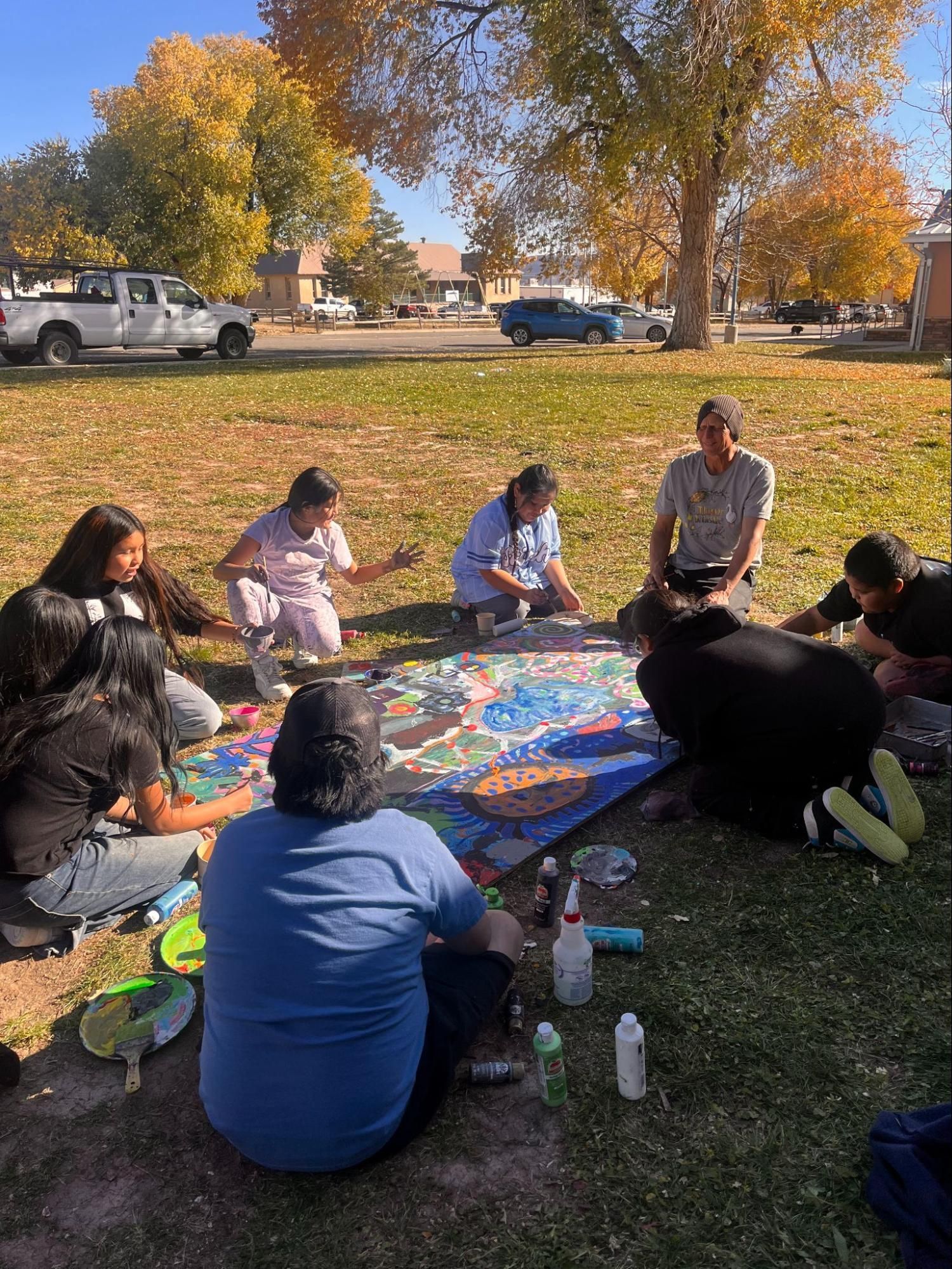People sitting in a circle outdoors on grass, gathered around a colorful blanket under autumn trees.