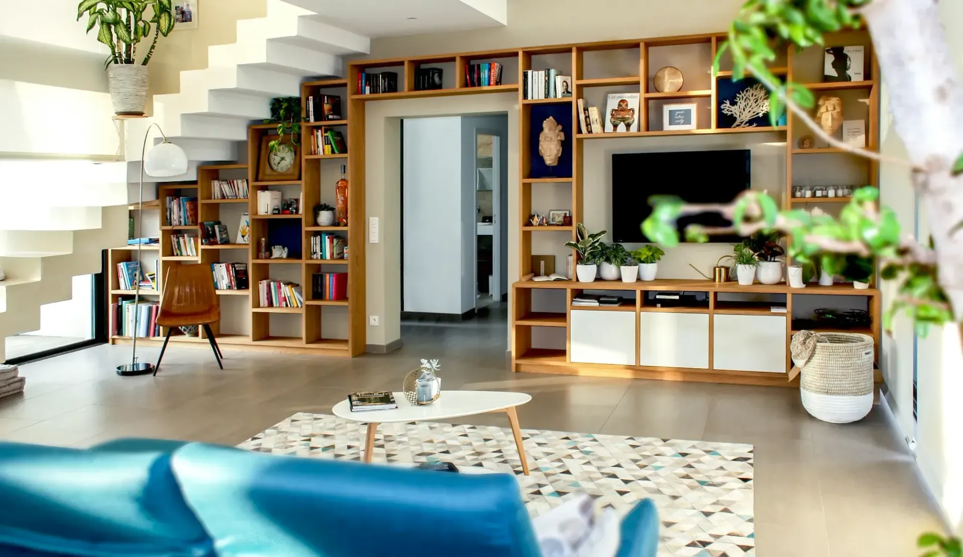 A living room with a blue couch , stairs , shelves and a flat screen tv.