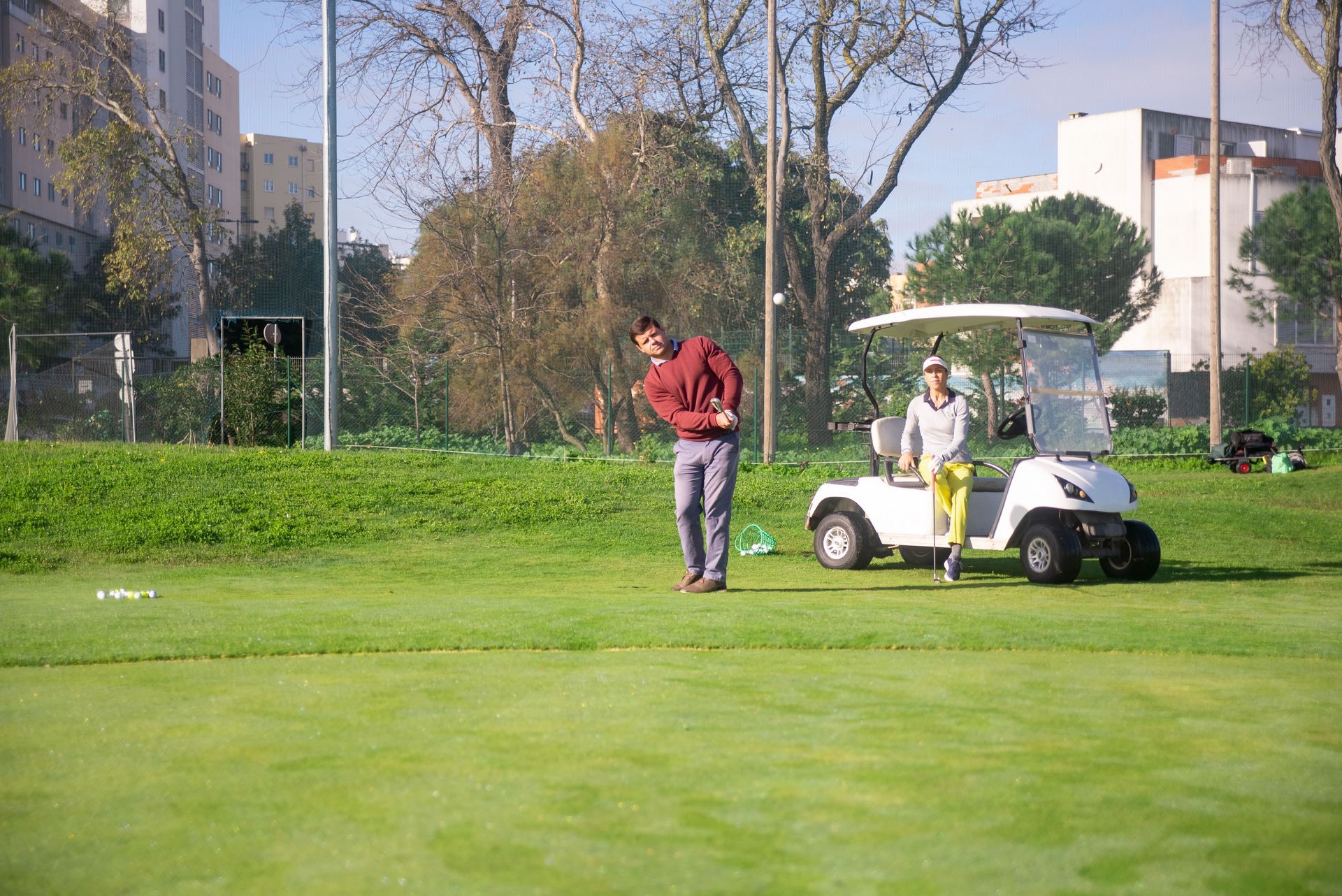 Two people beside a white golf cart on a green course with trees and buildings in the background