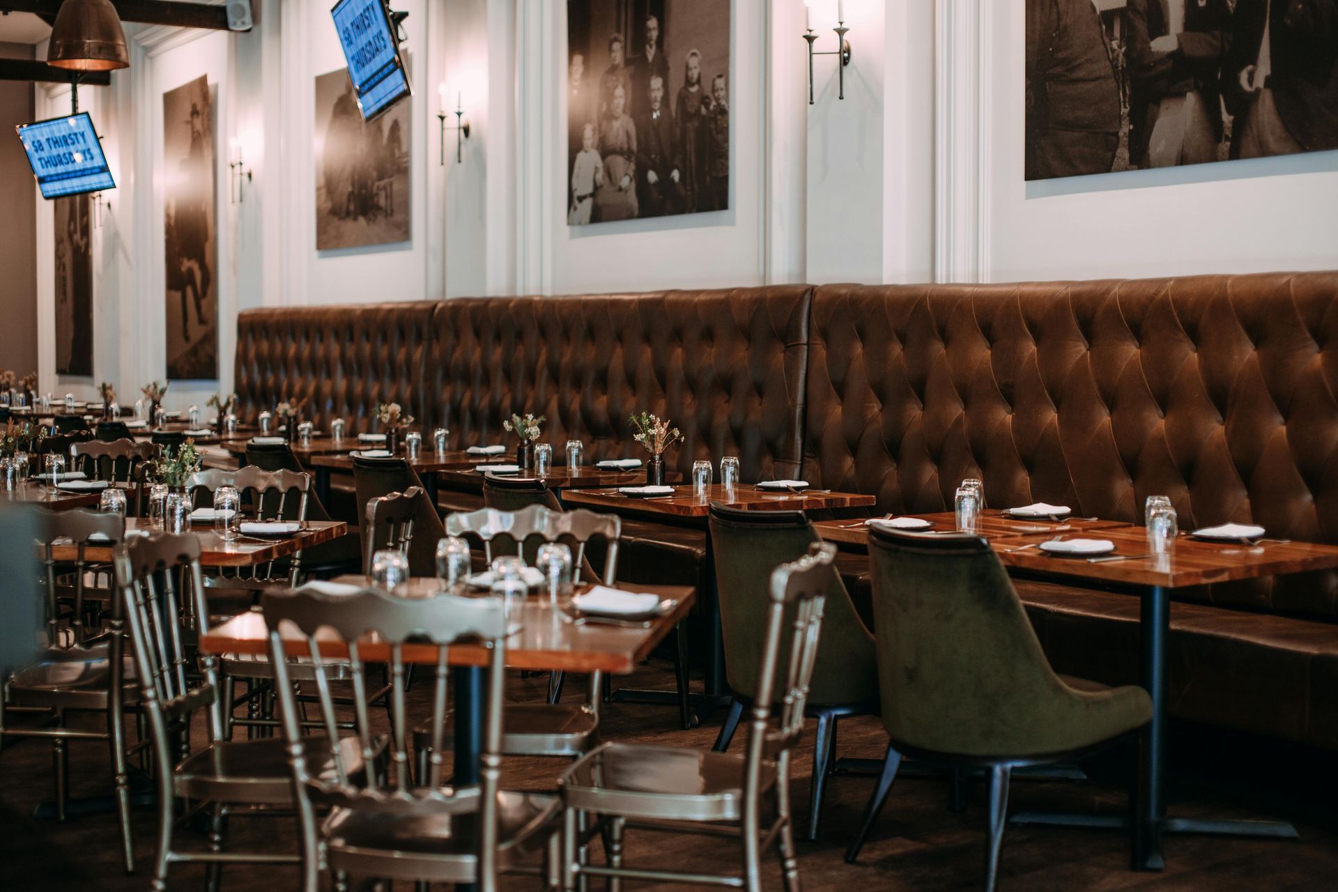 Empty upscale restaurant dining room with tufted brown banquette seating and set tables beneath framed photos
