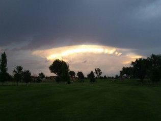 Stormy sky over a green field with dark trees and a bright break in the clouds at sunset