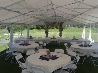White tent reception setup with round tables, white chairs, and floral centerpieces on a grassy lawn.
