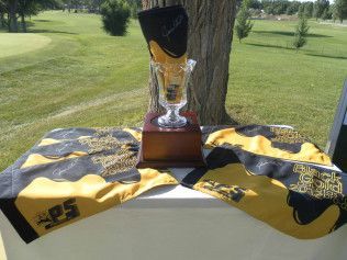 Trophy on a table outdoors, surrounded by black-and-gold sports scarves or sashes in a grassy field.