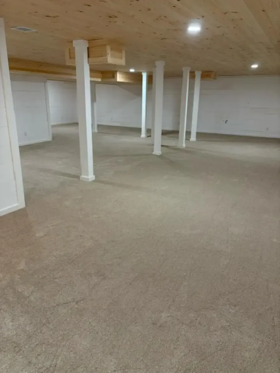 Empty carpeted basement with white support columns and a low wood ceiling