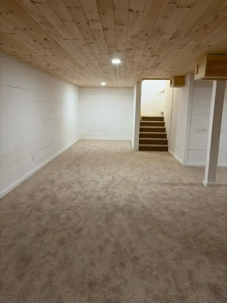 Empty carpeted room with white walls, wood ceiling, and stairs leading to a door on the right.