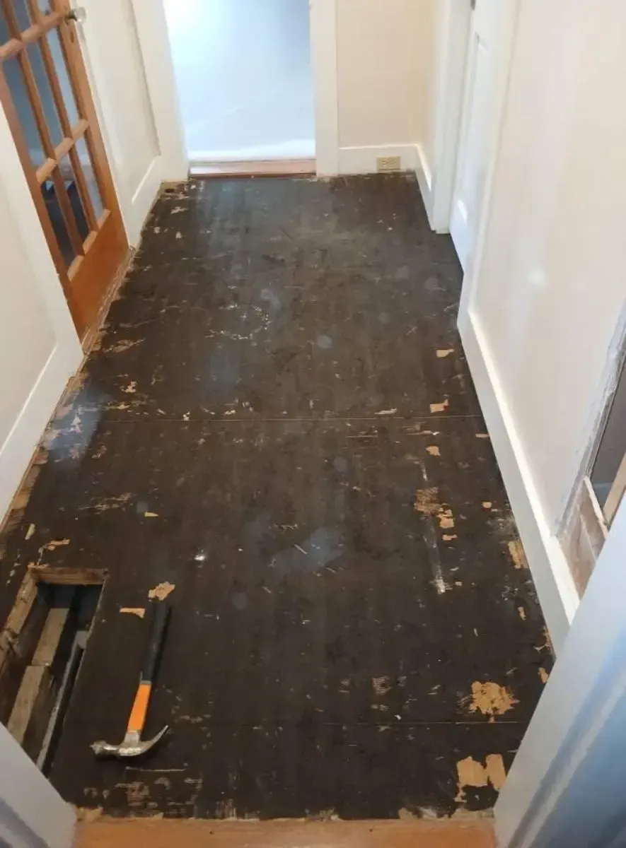 Hallway with damaged dark flooring, hammer visible, doorway at each end.