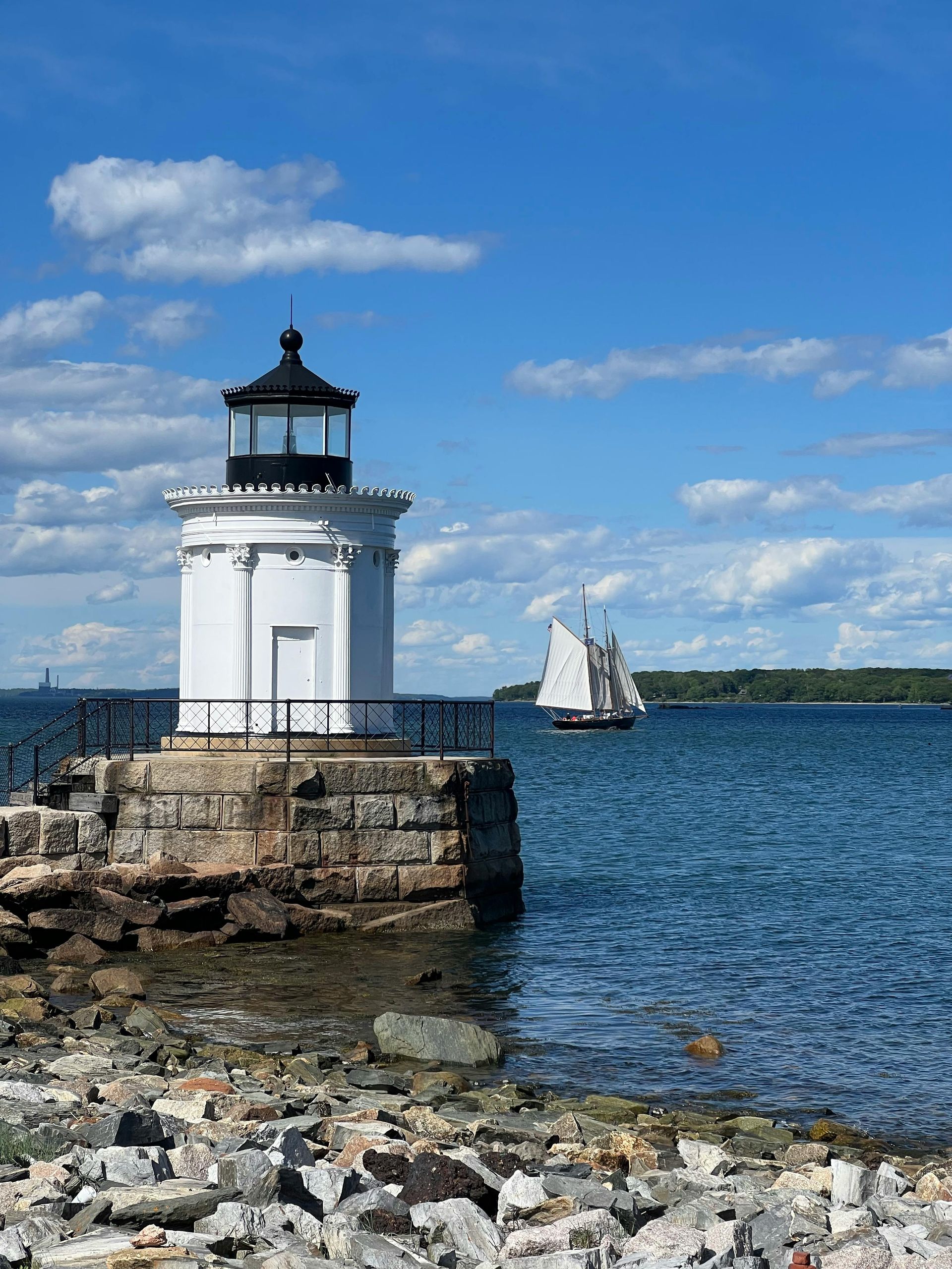 A lighthouse on a rocky shoreline with a sailboat in the background