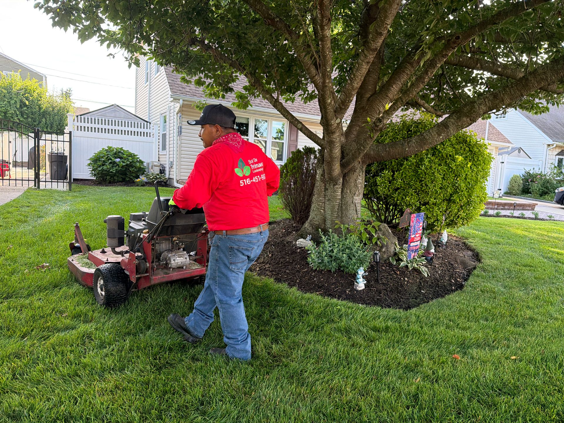 Worker in red shirt using a lawn mower near a tree in a manicured suburban yard