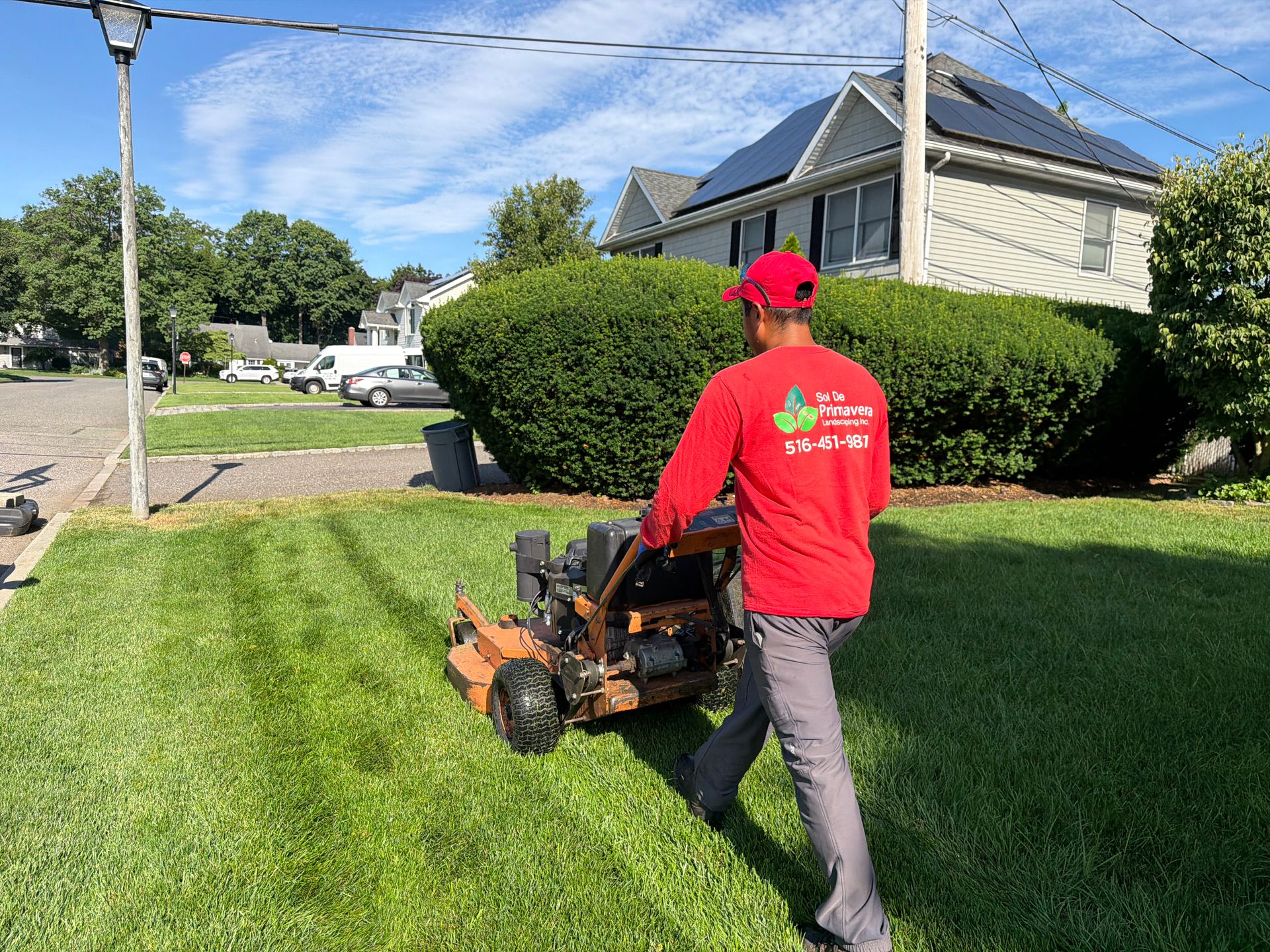 Person in red shirt mowing a grassy lawn in a suburban yard on a sunny day