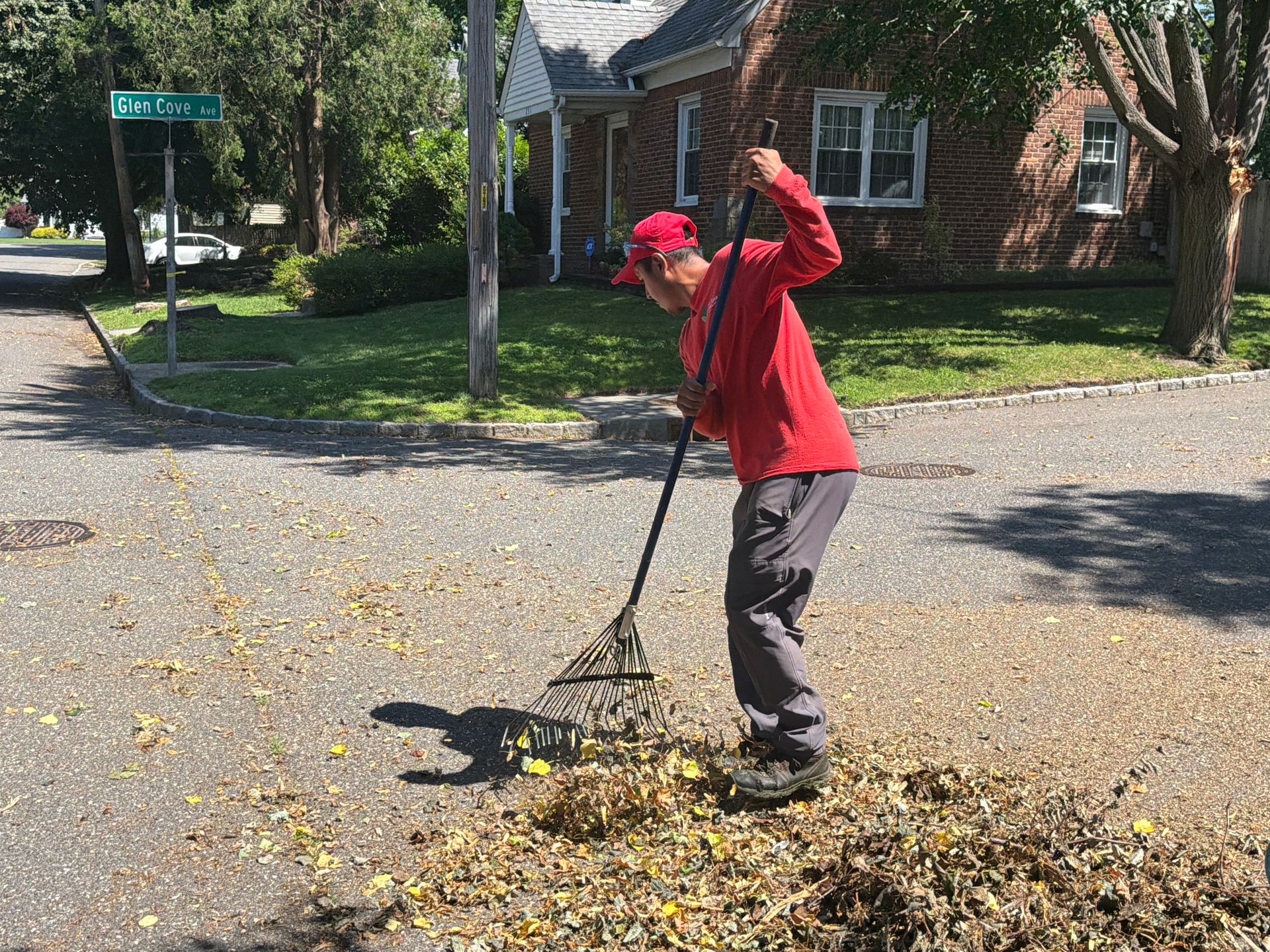 Person in red raking leaves on a sunny residential street corner