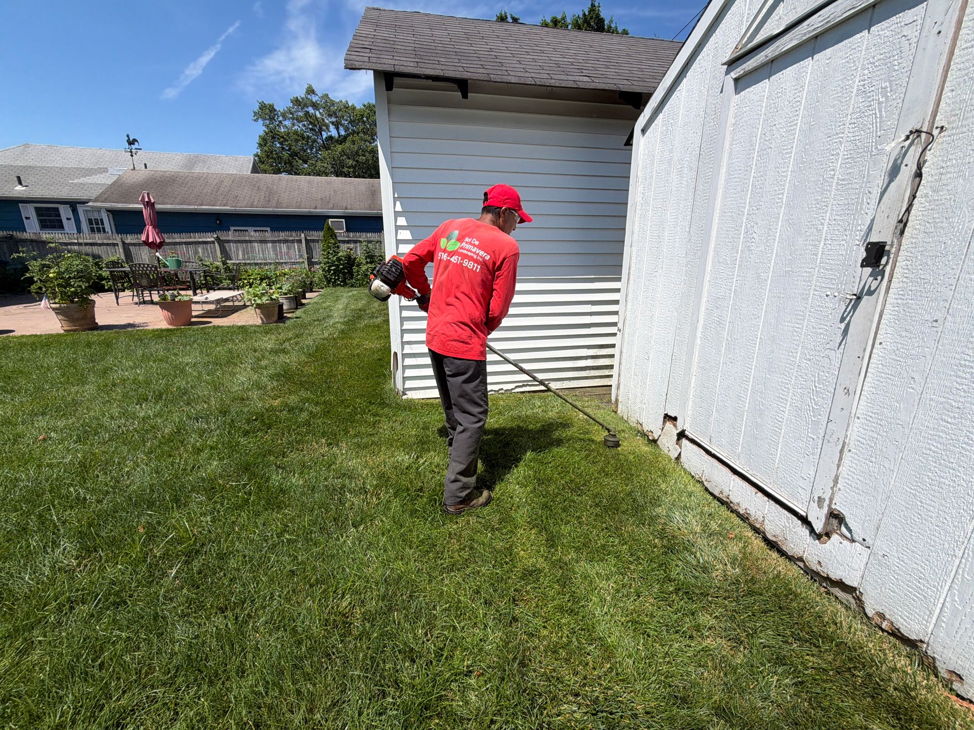 Person in red shirt using a string trimmer along a white shed beside a grassy yard.