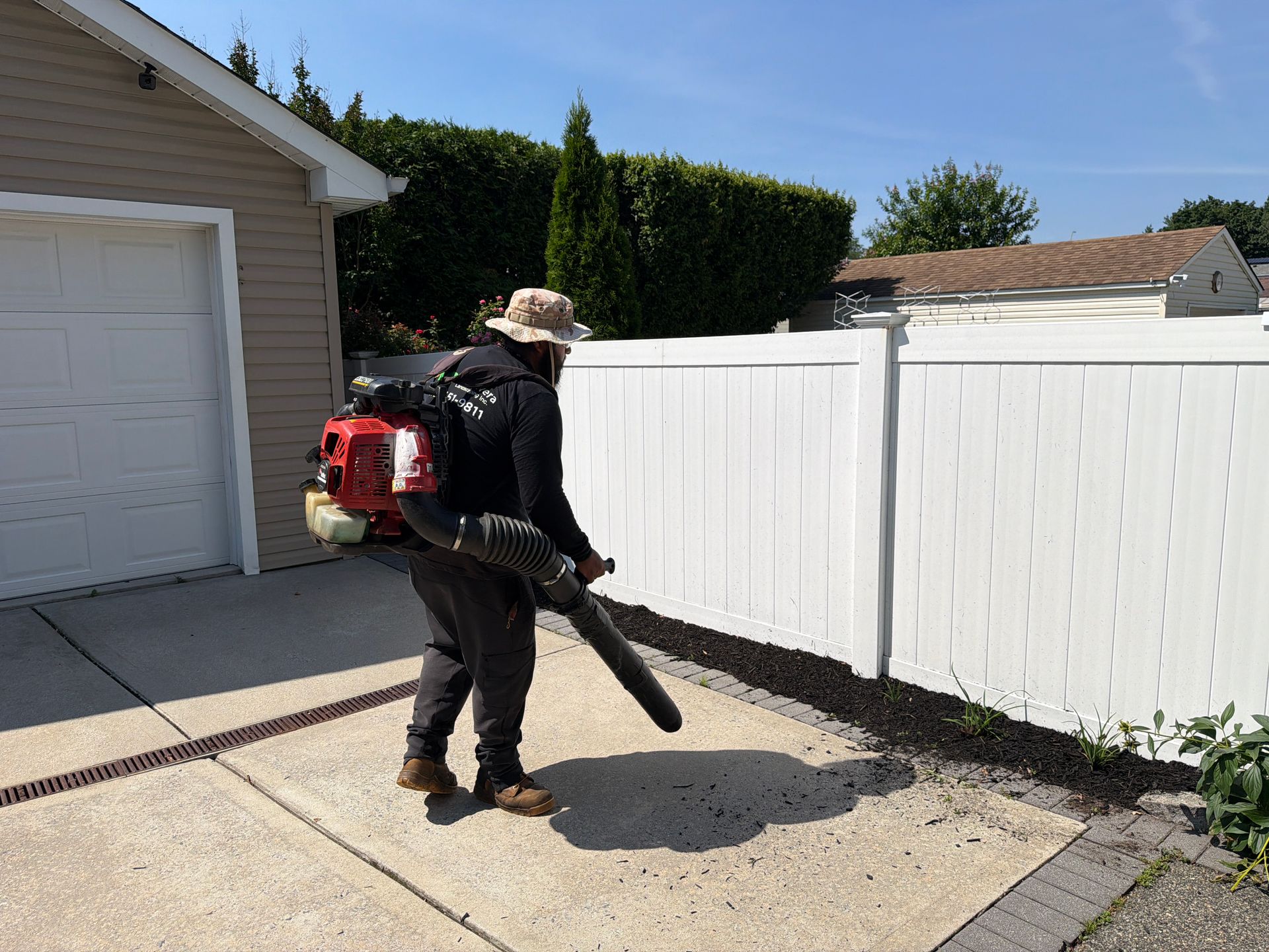 Person using a backpack leaf blower to clear a driveway beside a white fence.