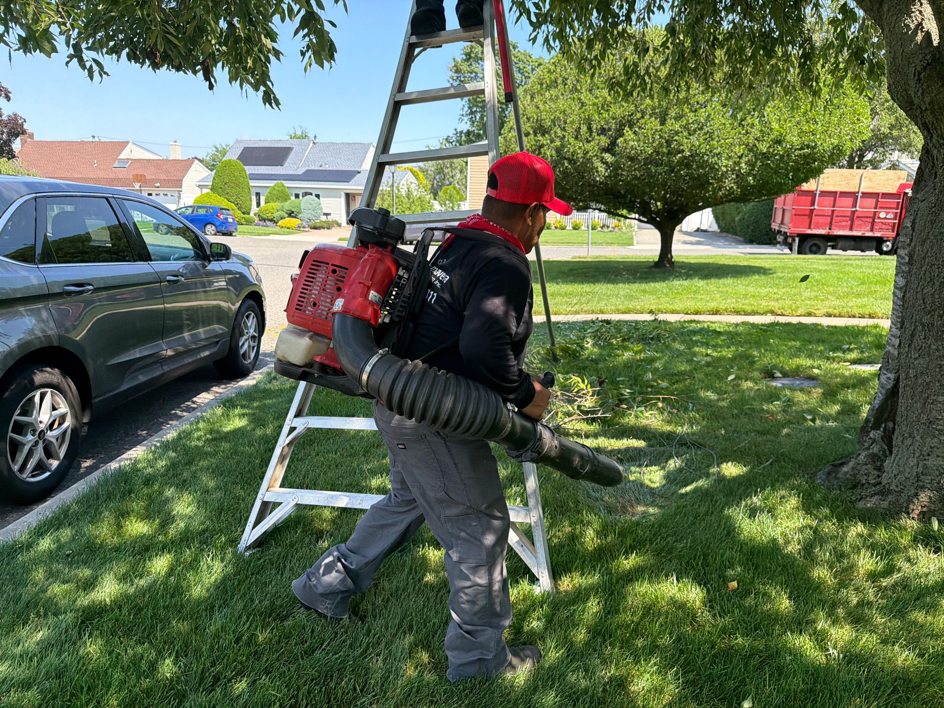 Worker with backpack leaf blower standing by a ladder on a grassy yard near parked cars and trees.