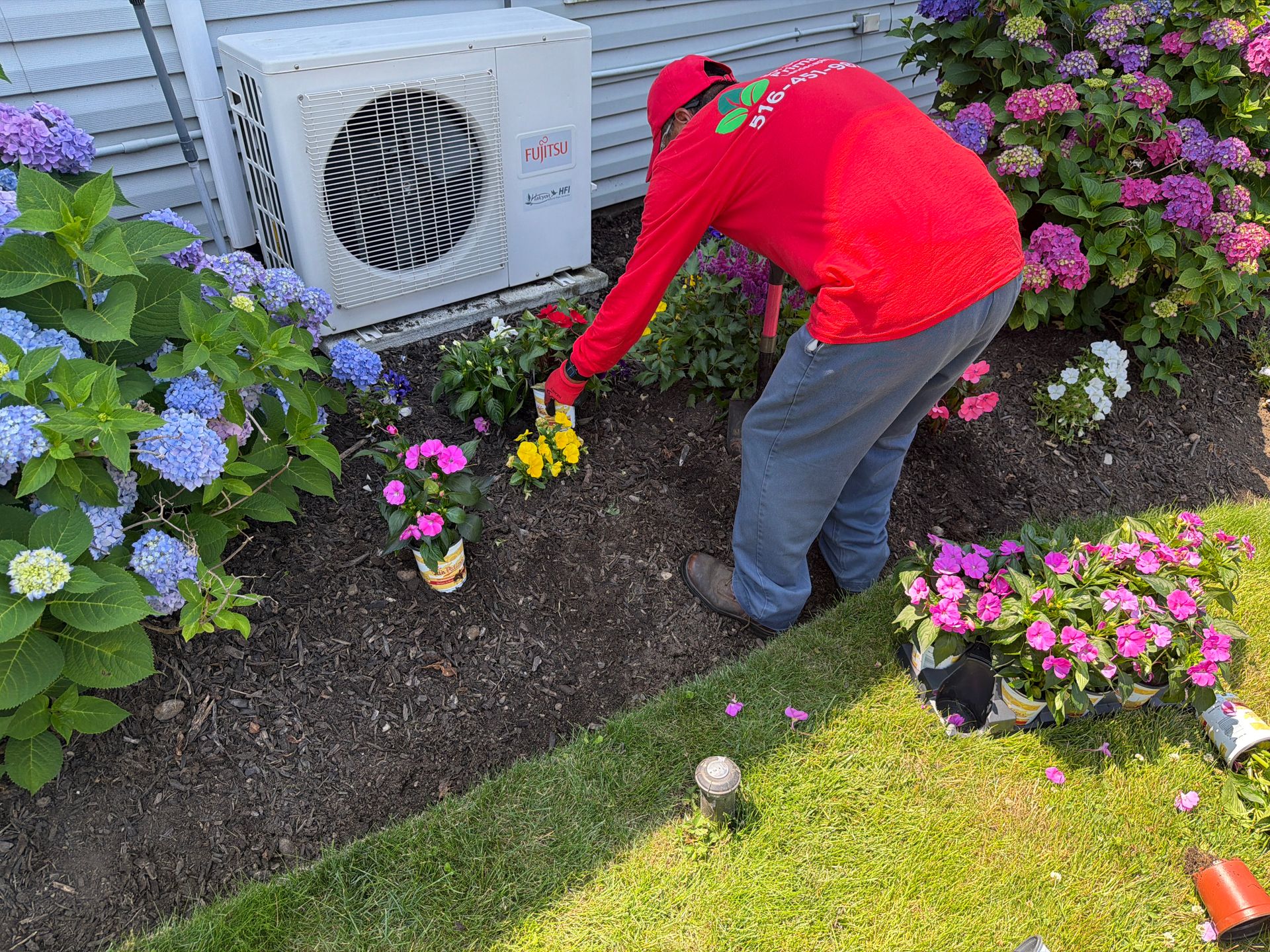 Person in a red jacket tending pink and yellow flowers beside a house air conditioner.