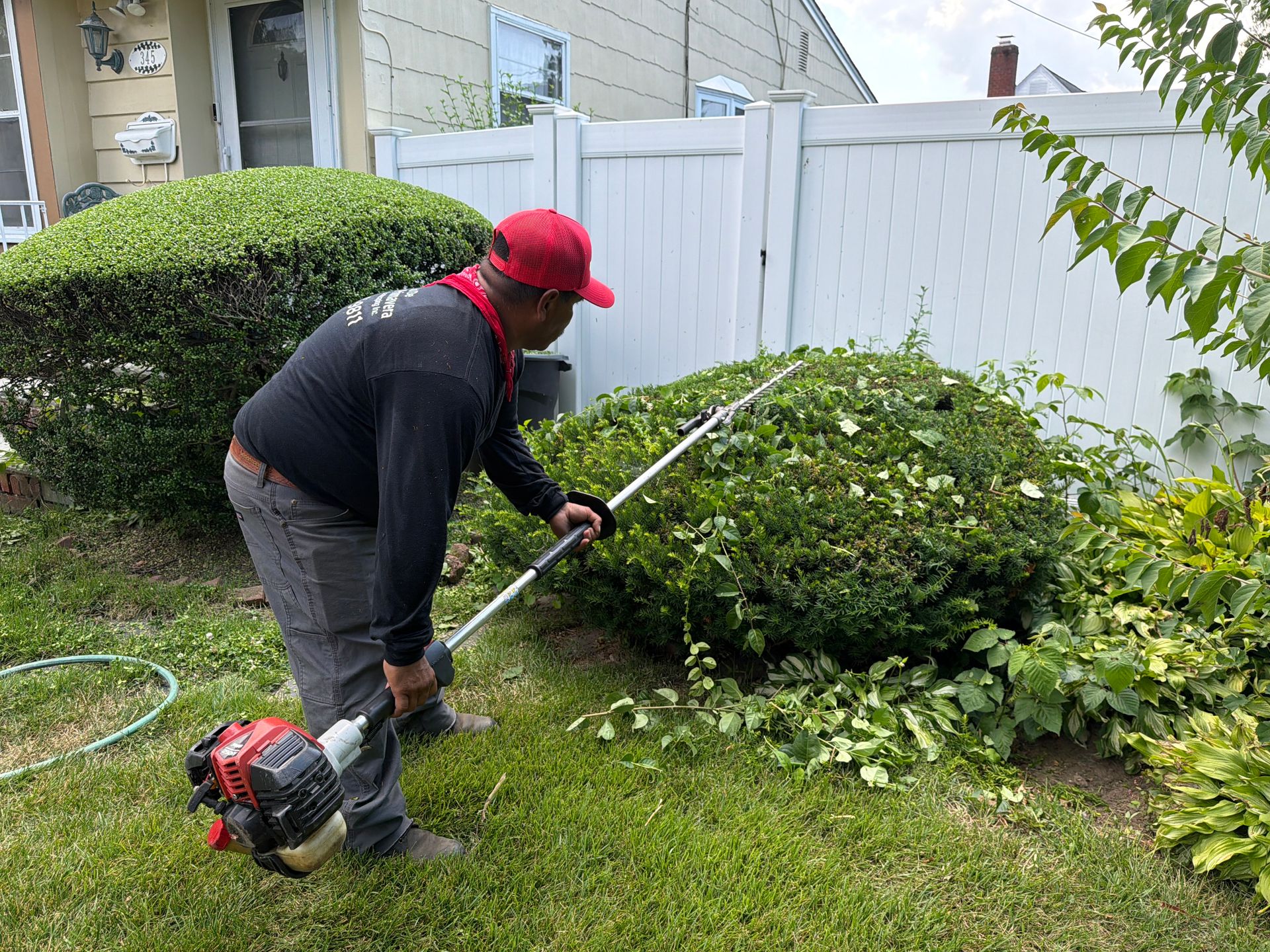 Worker using a gas-powered trimmer to cut shrubs along a white fence in a backyard