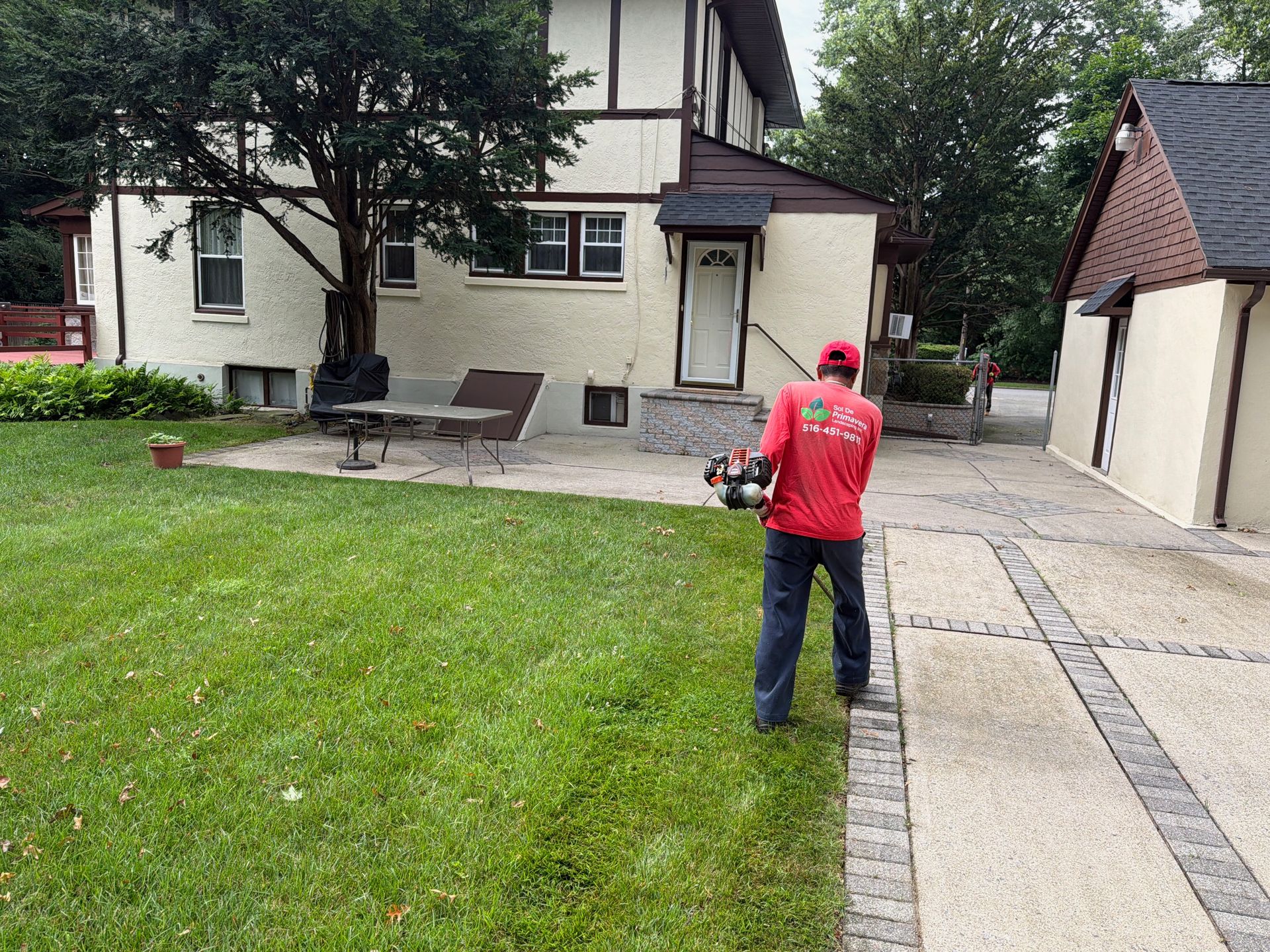 Person in a red shirt mowing a lawn beside a house and paved walkway.