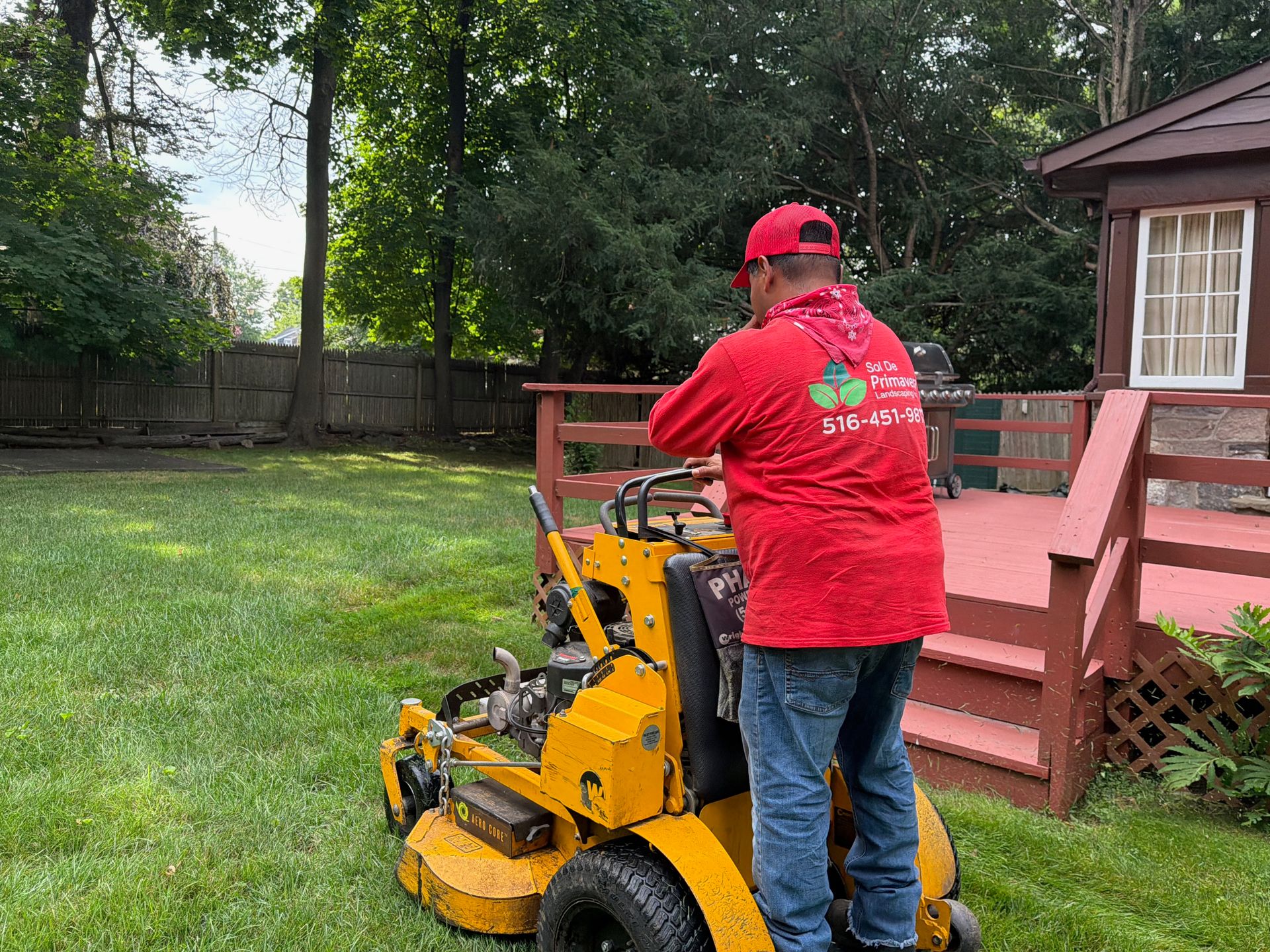 Person in red shirt and cap using a yellow mower beside a red deck in a backyard