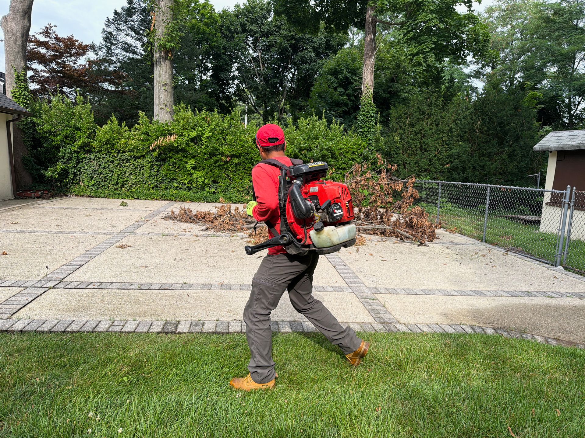 Person in red cap and backpack walking along a driveway beside a grassy yard and trees.