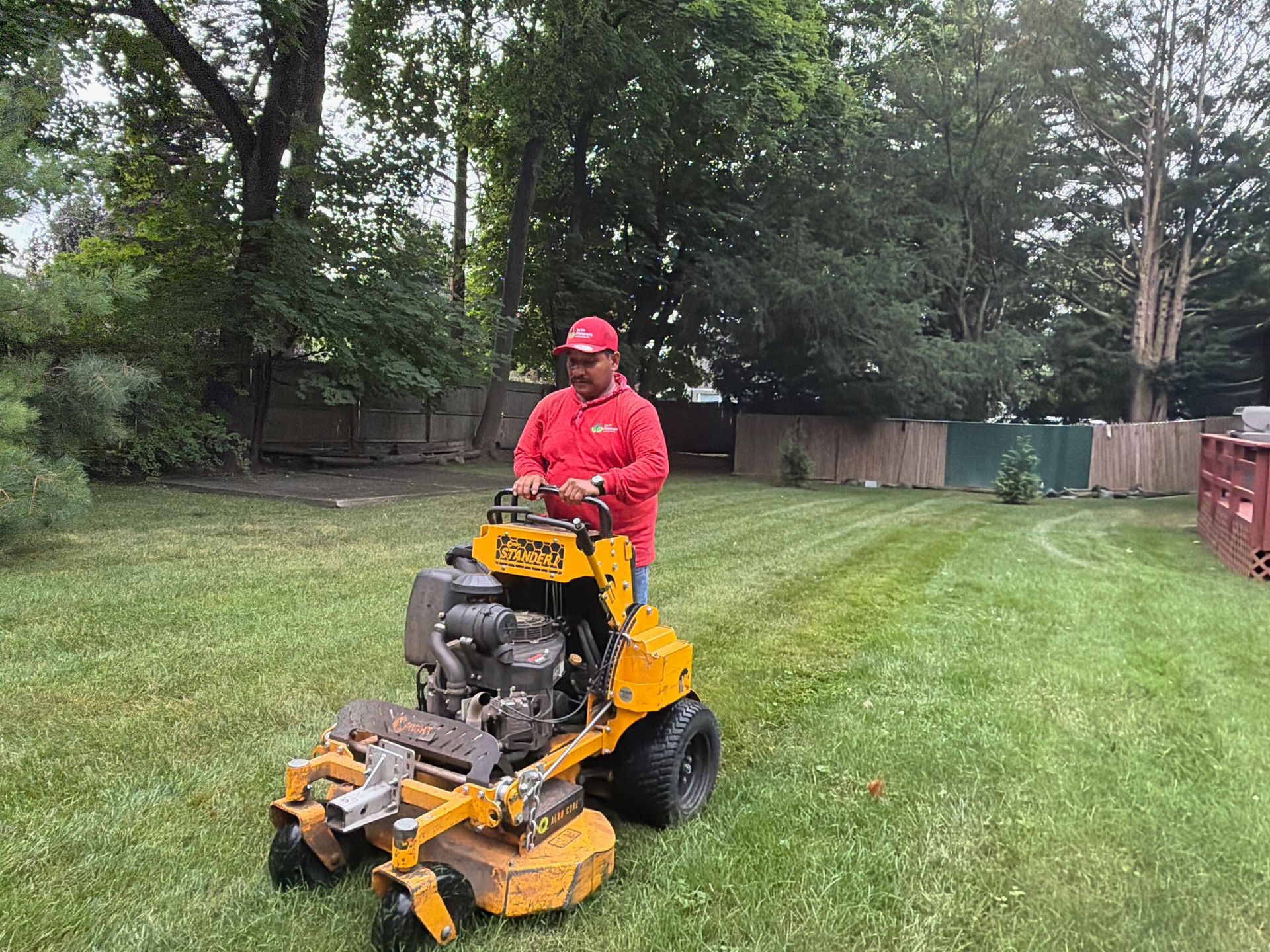 Man in red riding a yellow lawn mower on a grassy yard with trees and a fence nearby
