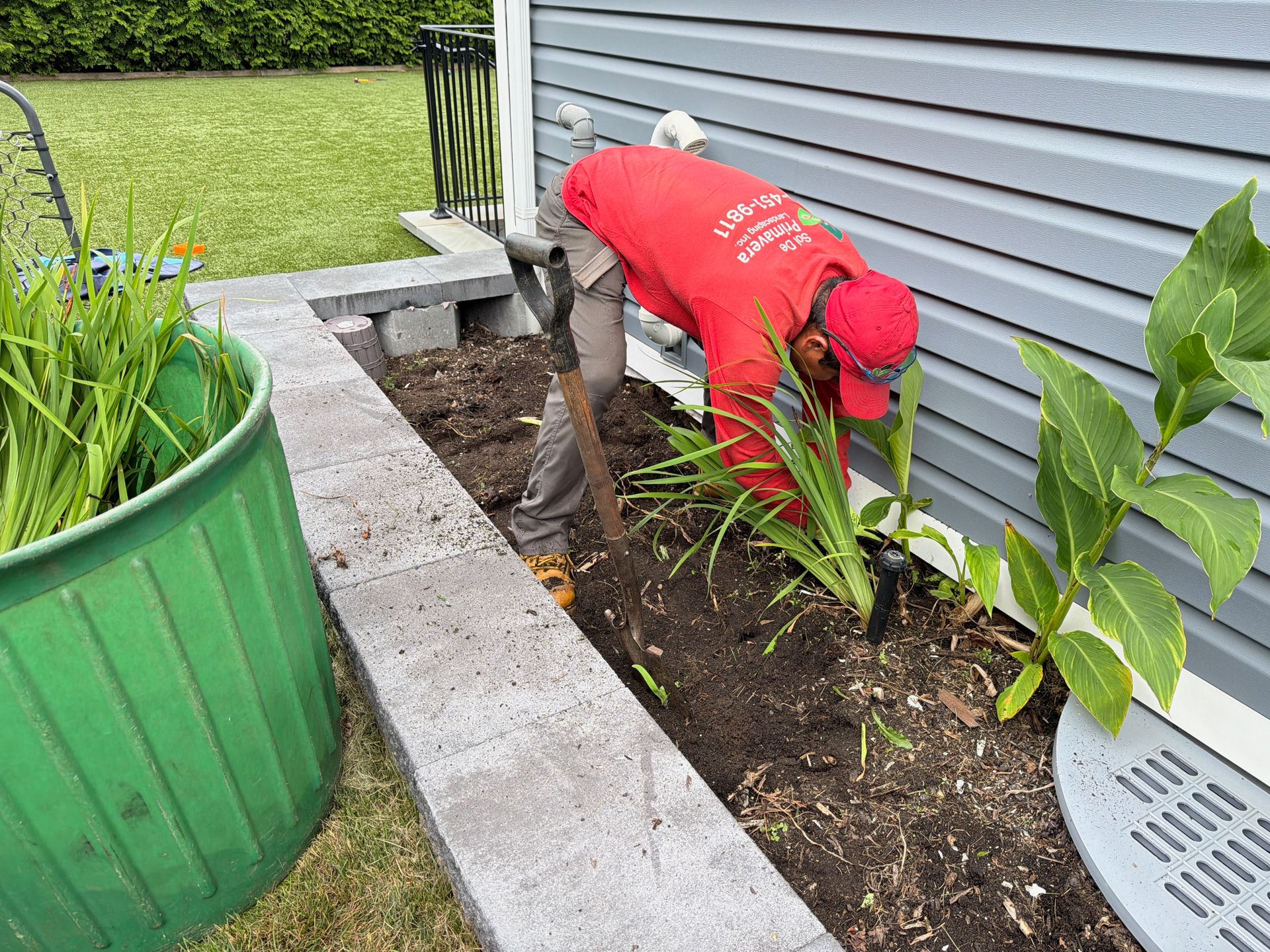 Person in a red shirt kneels by a house foundation, digging soil beside a garden bed.