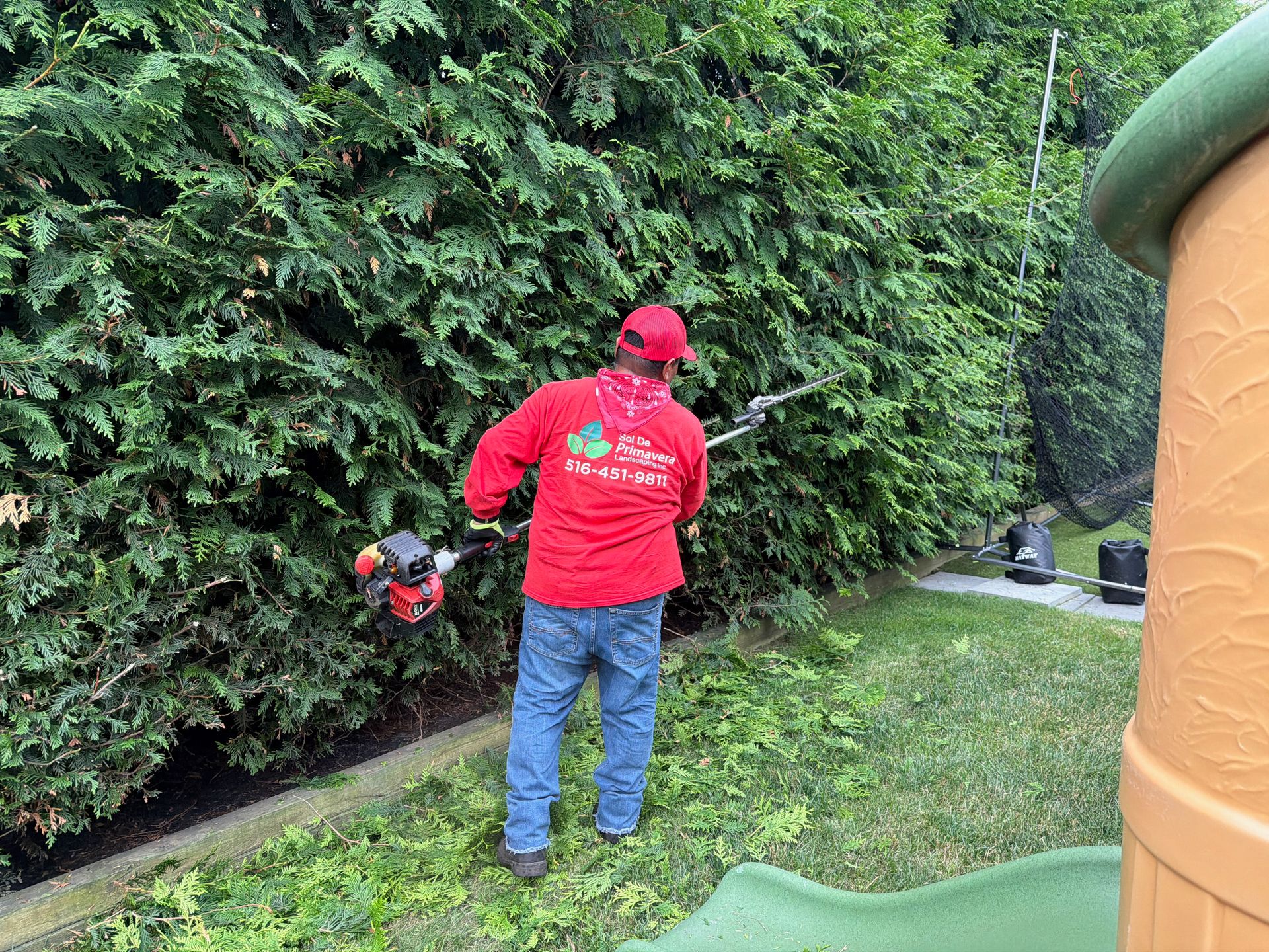 Person in red shirt and cap trimming a hedge in a backyard with a string trimmer