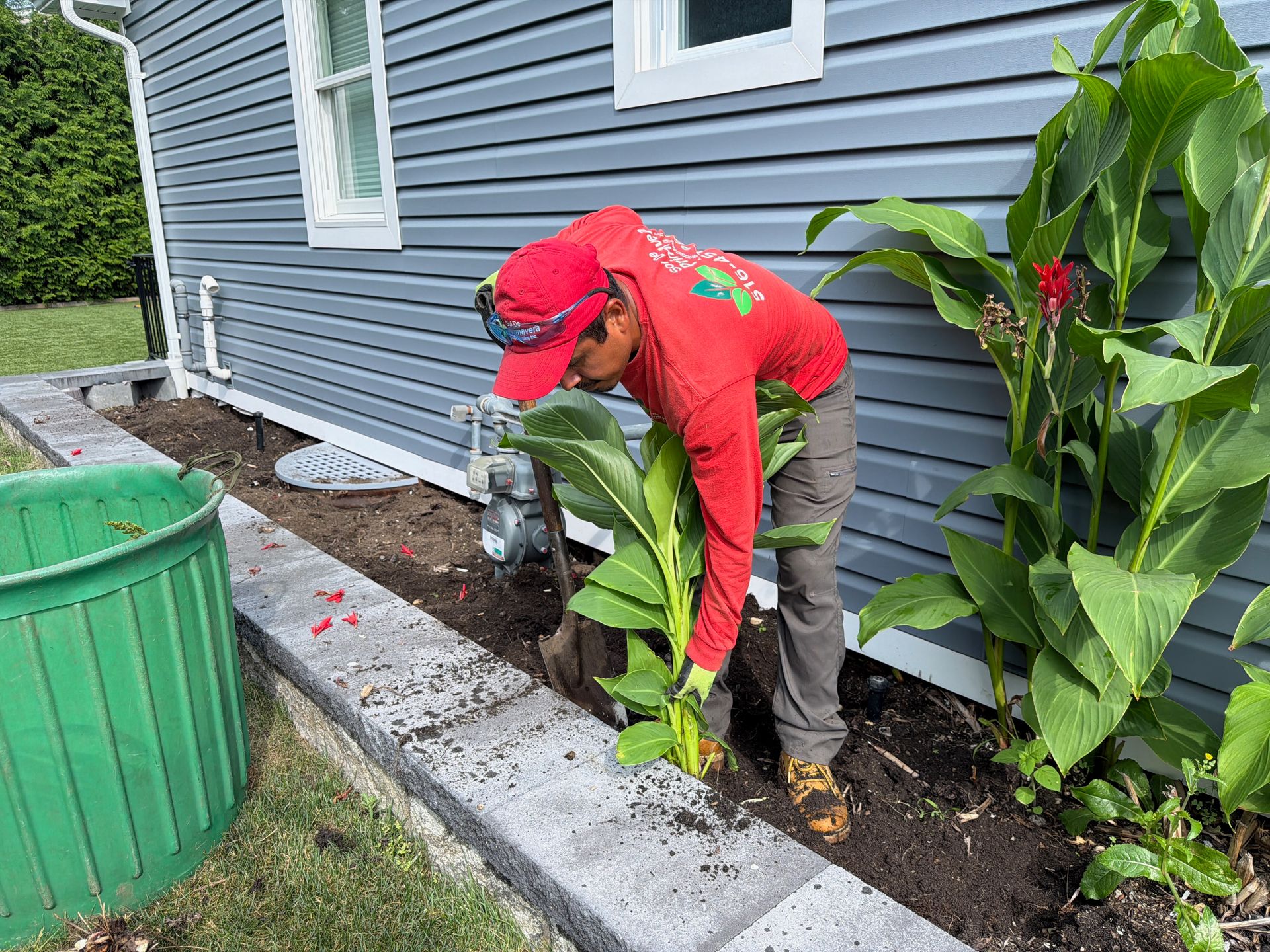 Person planting in a garden bed beside a blue house, next to a green trash bin.