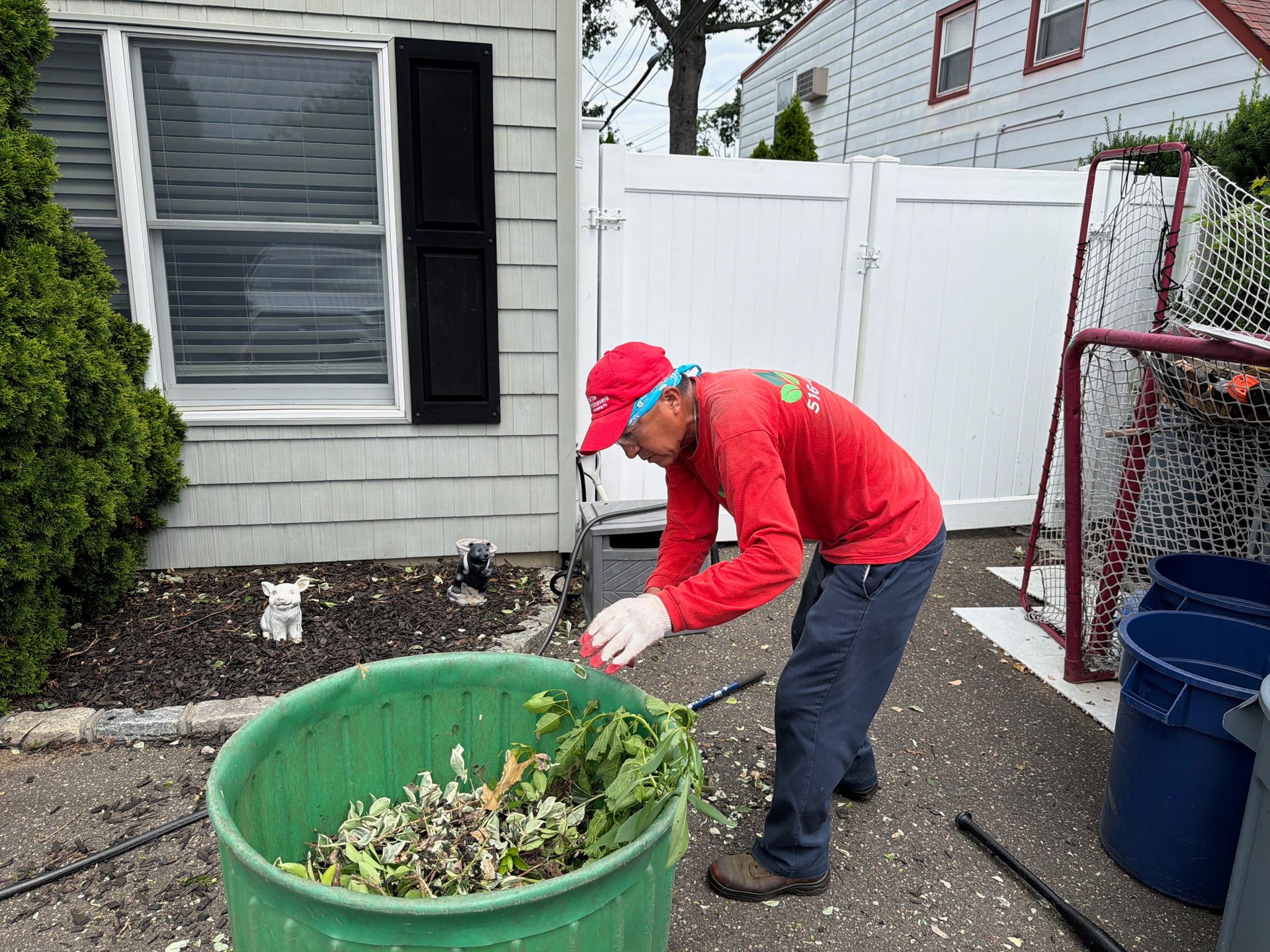 Person in red shirt and cap raking yard debris into a green bin beside a house.