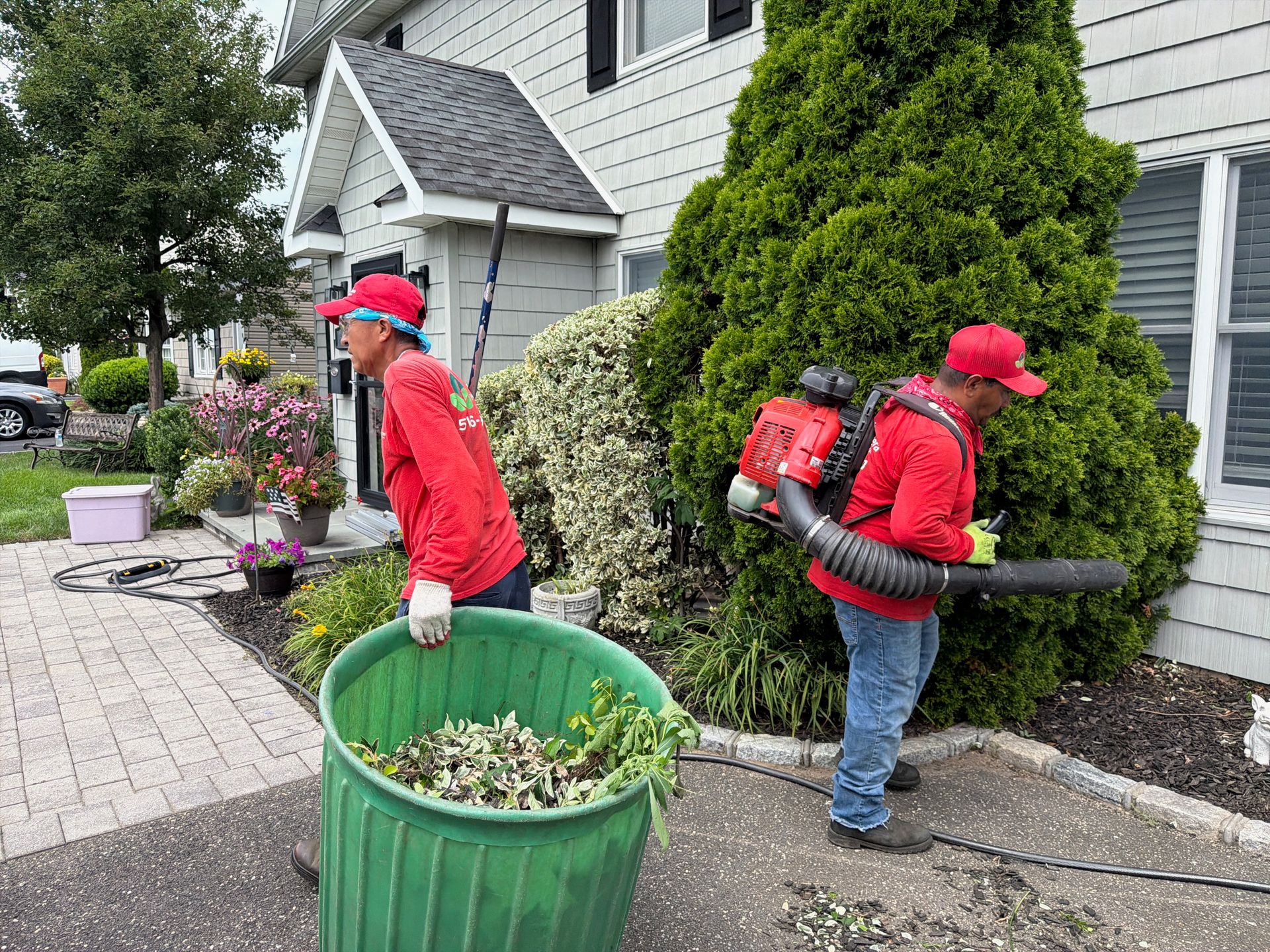Two workers in red shirts carrying leaf blowers and tubs along a sidewalk beside a house and shrubs